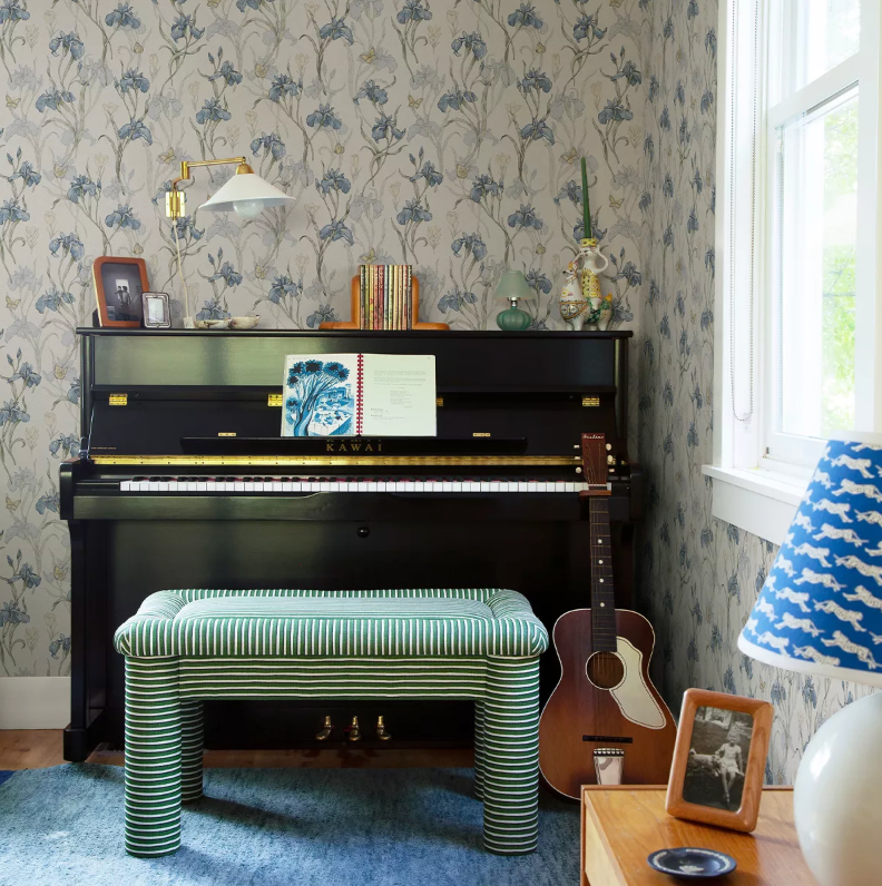 A black upright piano with a green striped bench, an acoustic guitar, and blue iris floral wallpaper in a cozy room.