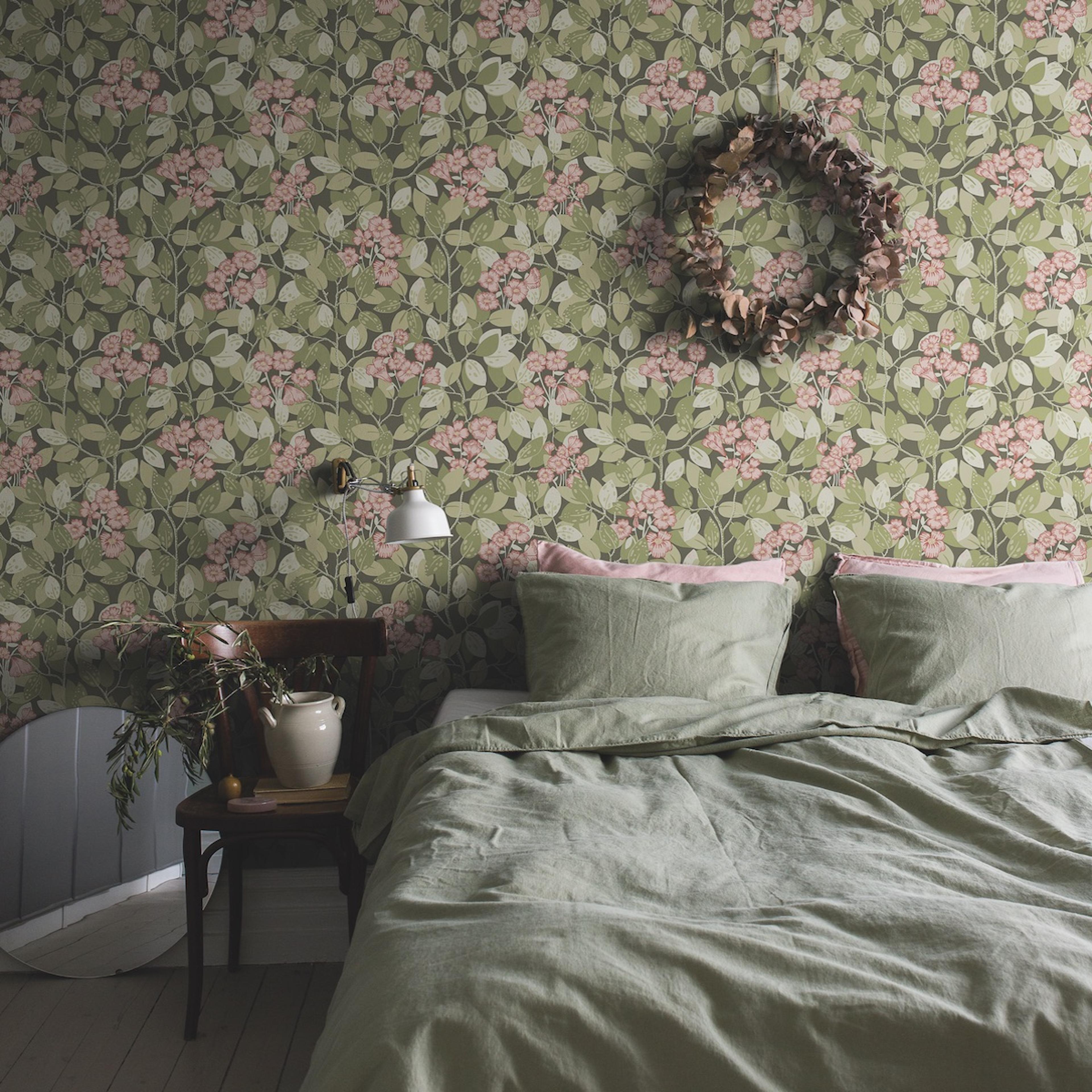 Bedroom with dark floral wallpaper featuring pink flowers and green leaves, a light green linen bed, and a dried wreath.