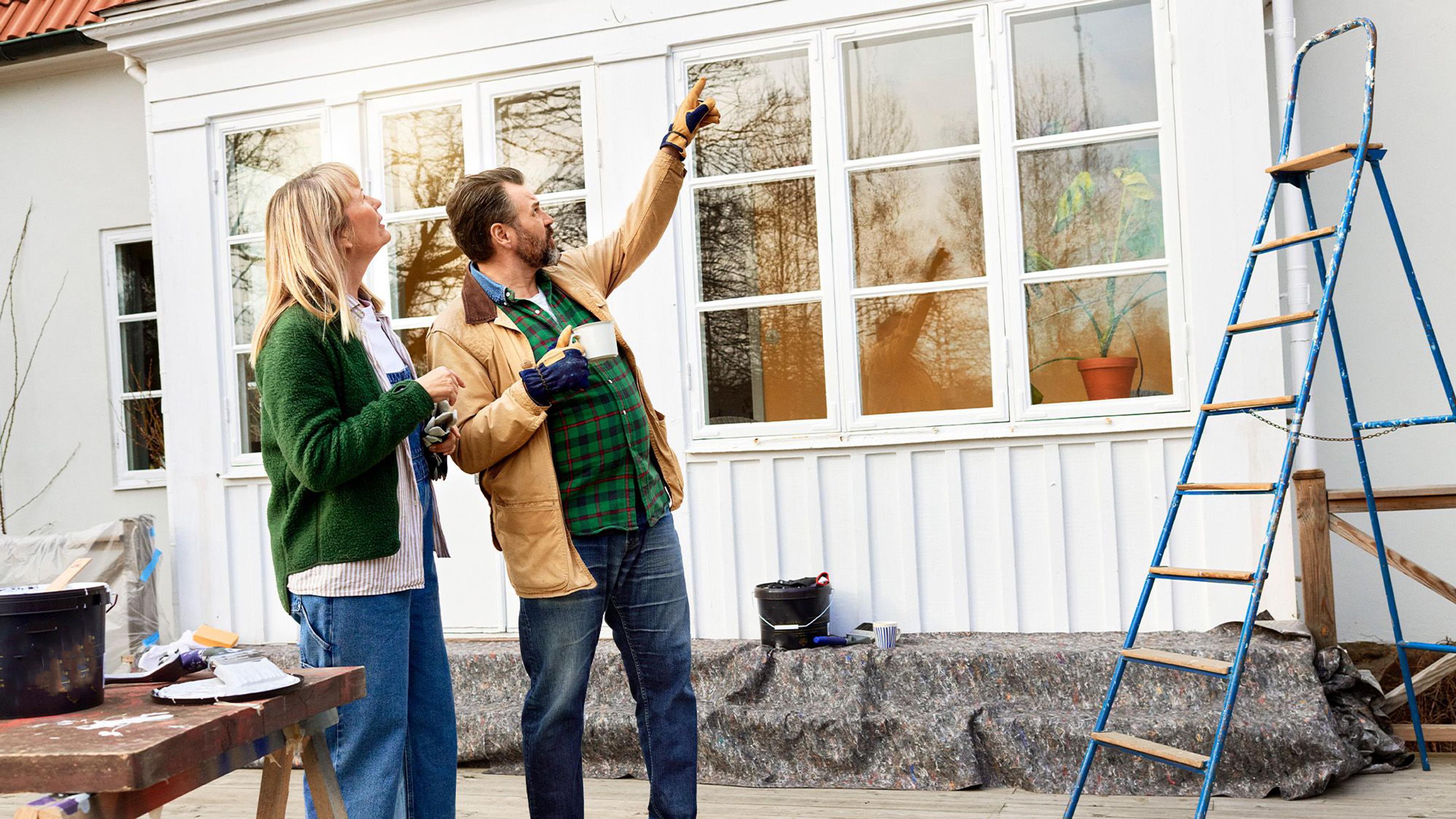 A man points to the white exterior of a house as a woman looks on; a ladder and renovation supplies are visible.