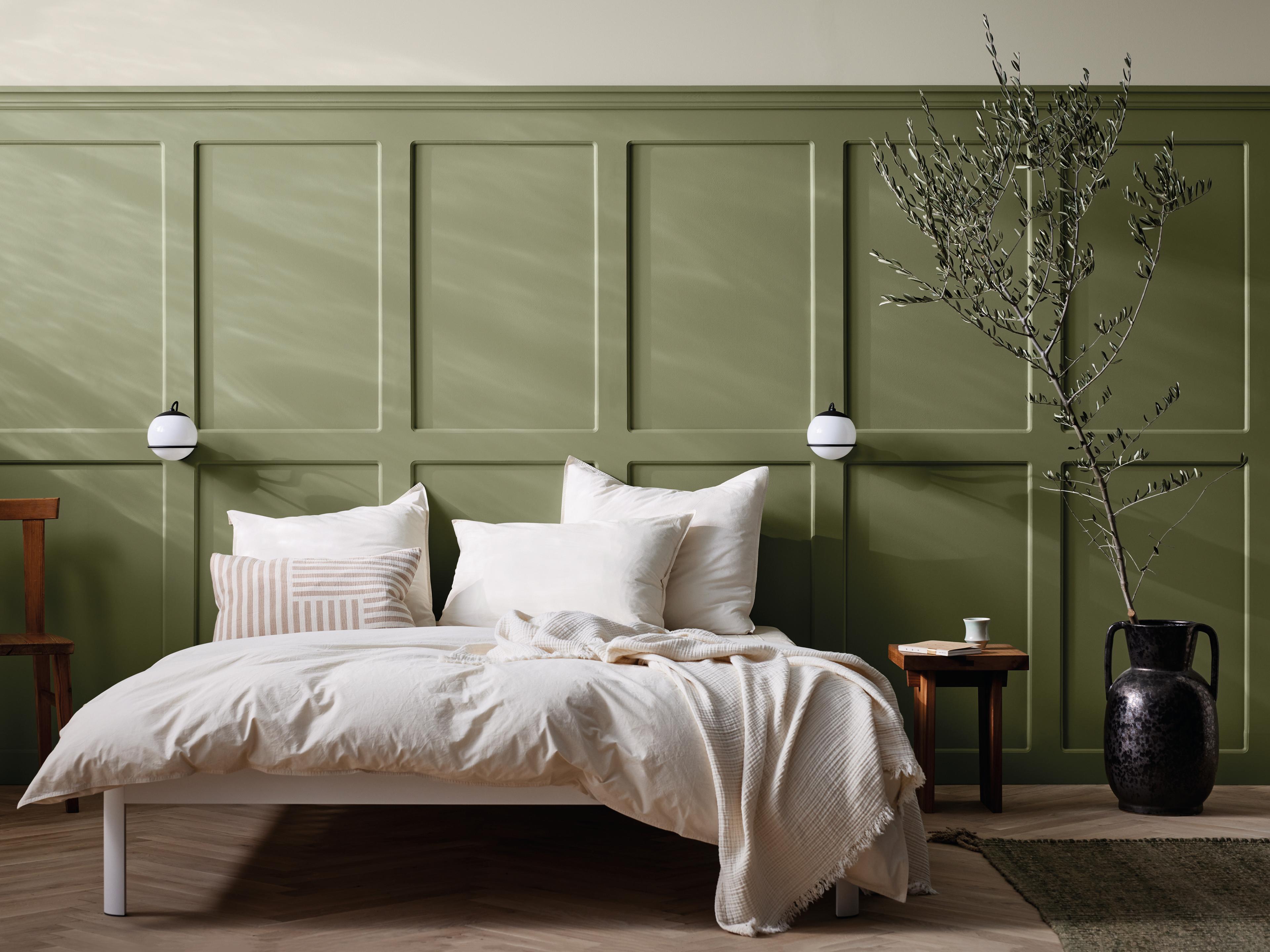 A bedroom with a white bed and cream bedding against a sage green paneled wall, featuring wall sconces and a large branch in a dark vase.