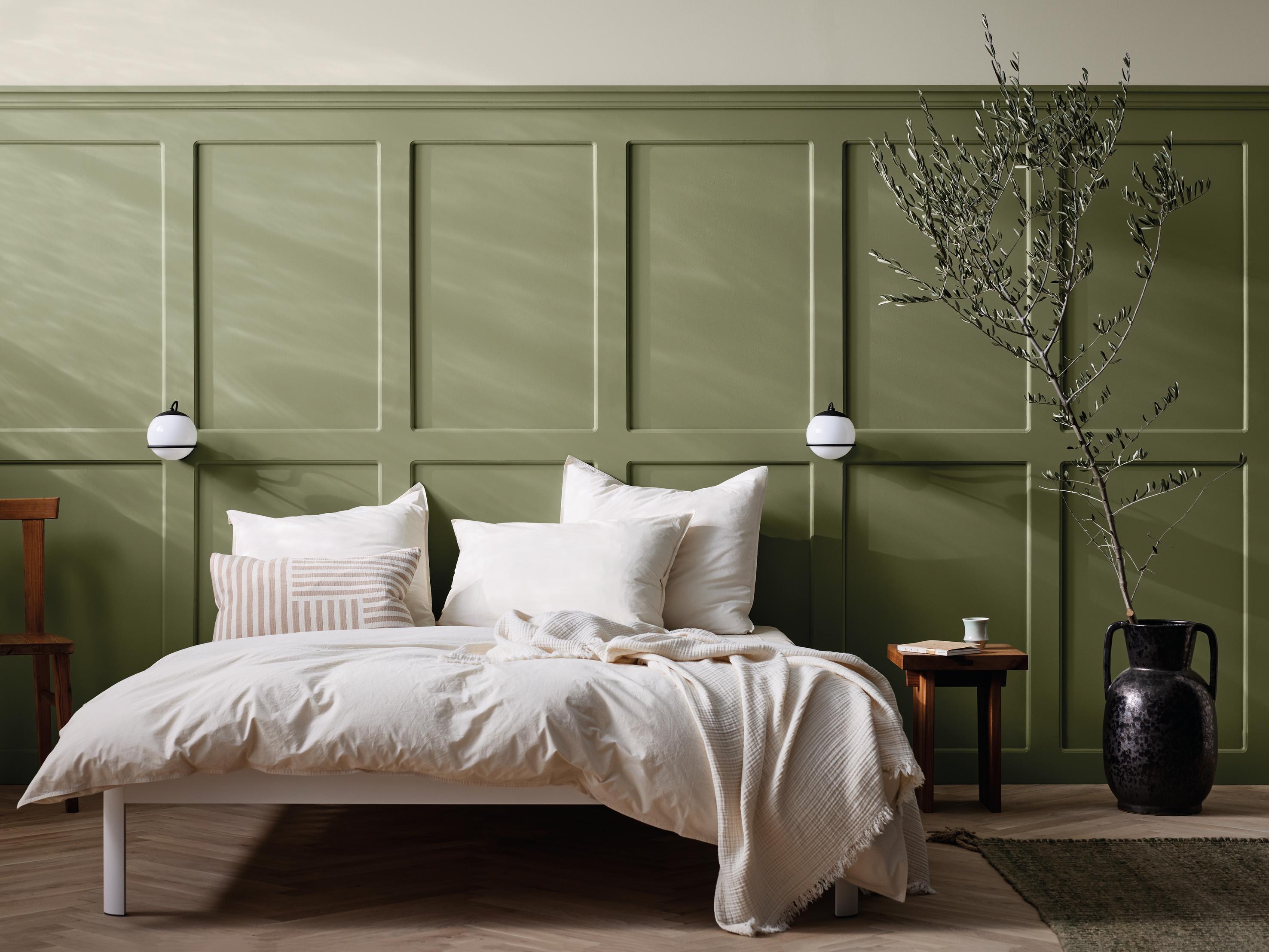 A bedroom with a white bed and cream bedding against a sage green paneled wall, featuring wall sconces and a large branch in a dark vase.