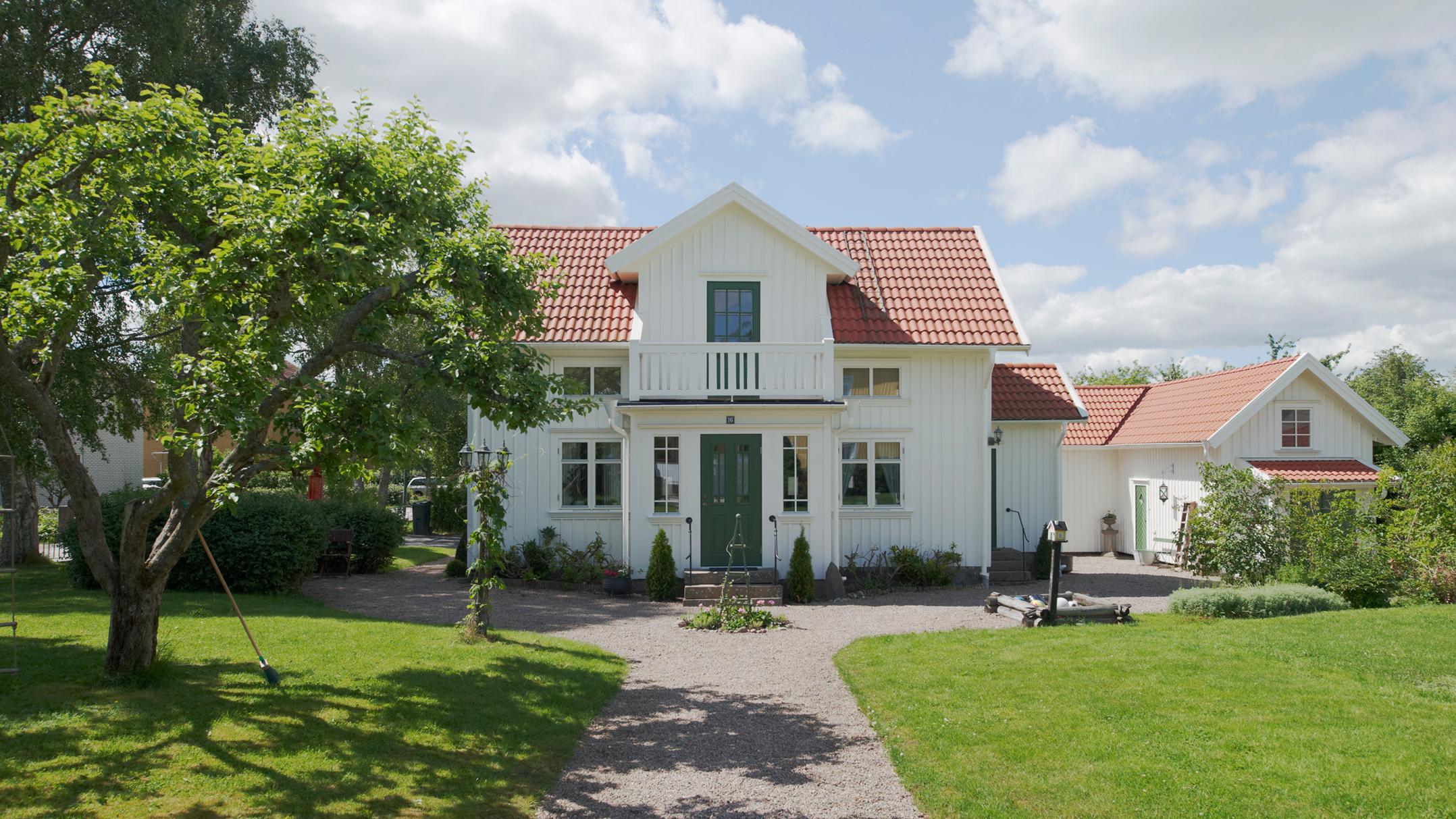 A white house with a red roof, green front doors, and a small balcony, set on a green lawn with a tree and gravel path under a sunny sky.