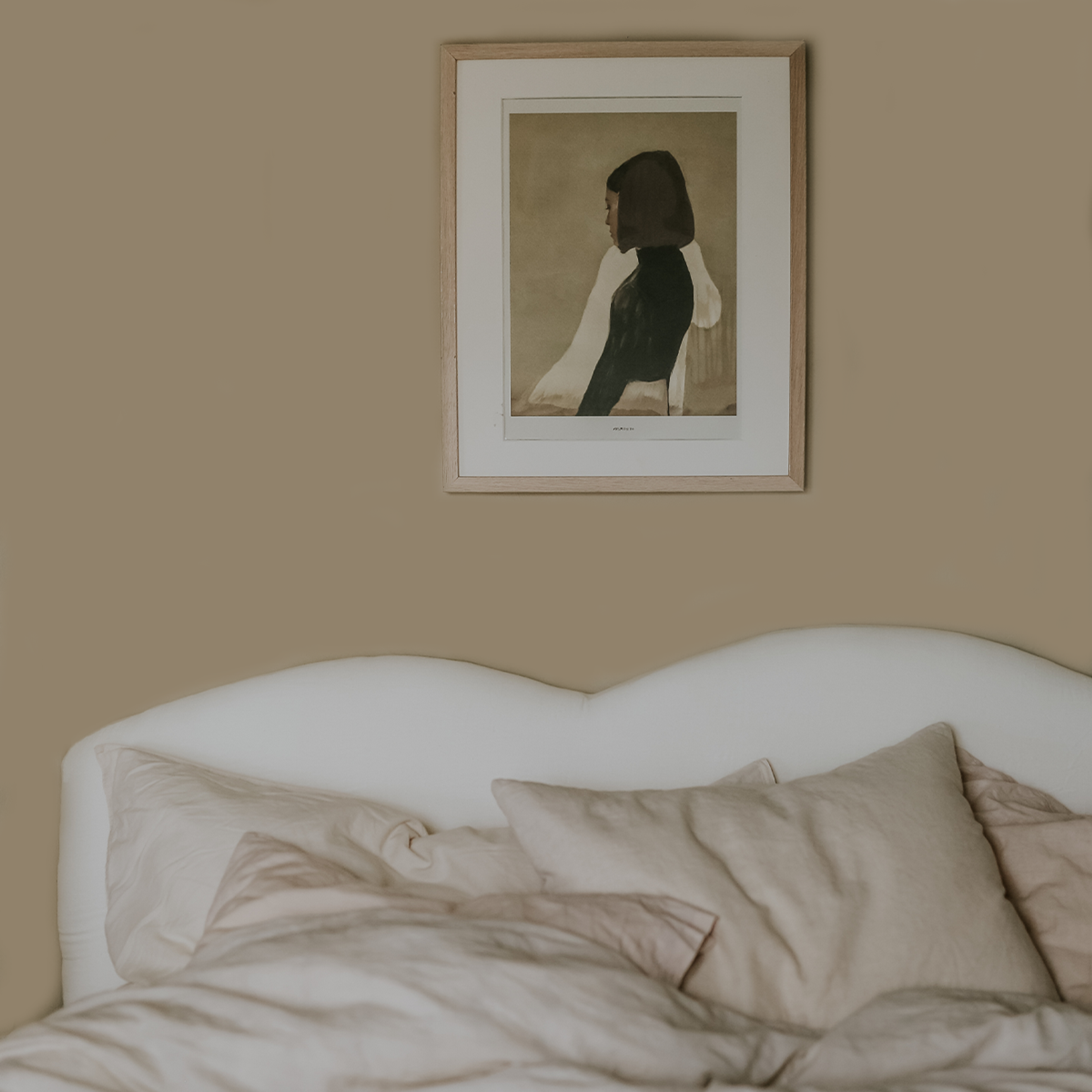 A framed art print of a woman in profile hangs on a tan wall above a white curved headboard and light-colored bedding.