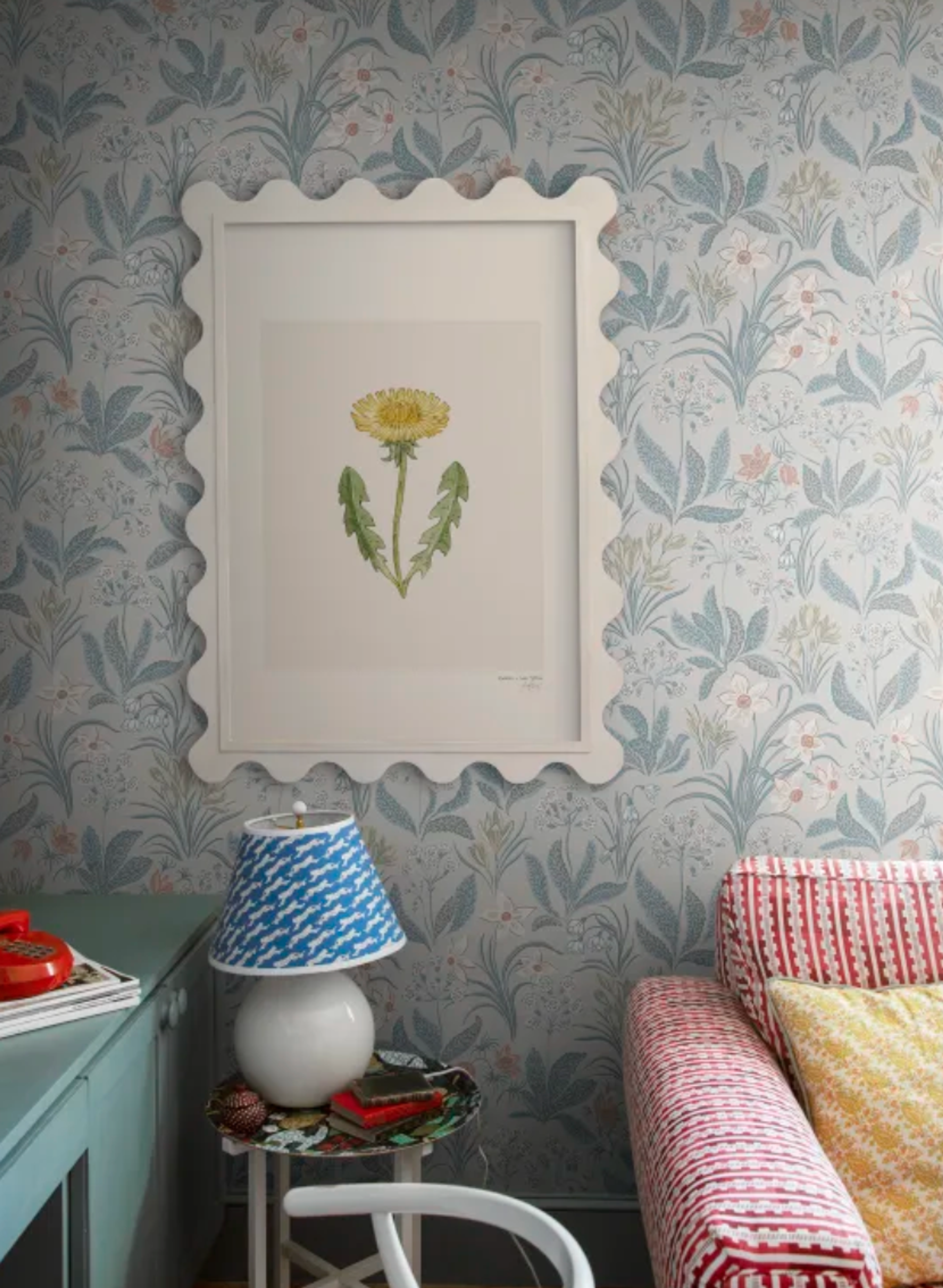A dandelion painting in a white scalloped frame on light blue floral wallpaper, with a lamp and striped sofa.