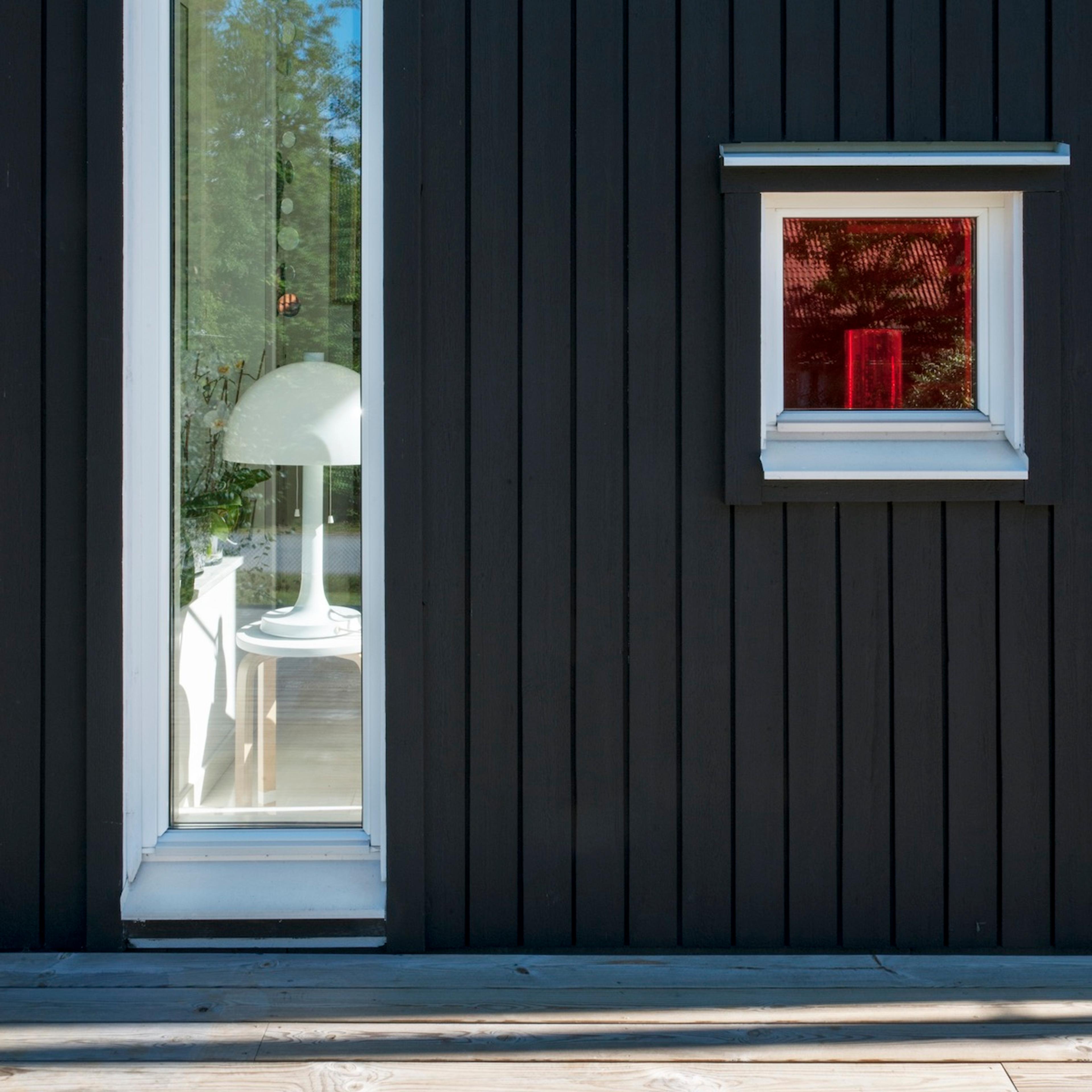 Dark slatted wall with a tall window showing a white lamp and a small window with a red reflection, above a wooden deck.