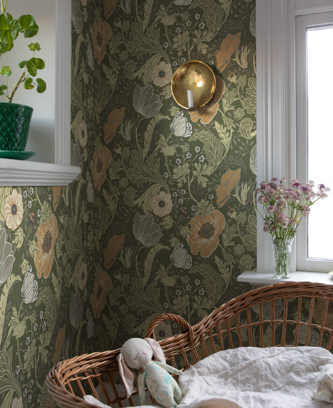 A room with dark green floral wallpaper, a brass sconce, and a wicker bassinet with a bunny toy.