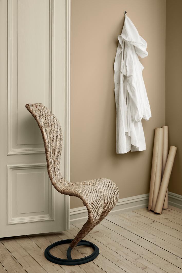 A cobra-shaped woven wicker chair with a black base on a light wood floor. A white shirt hangs on a tan wall.