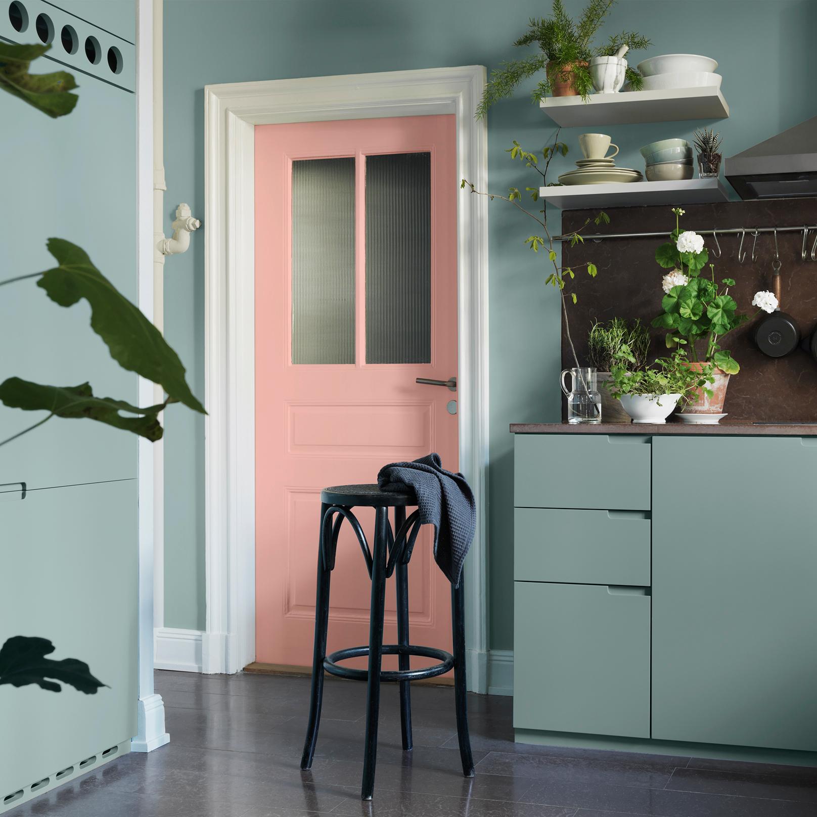 A kitchen with sage green cabinets, light blue walls, a pink door, a black stool, and potted plants.