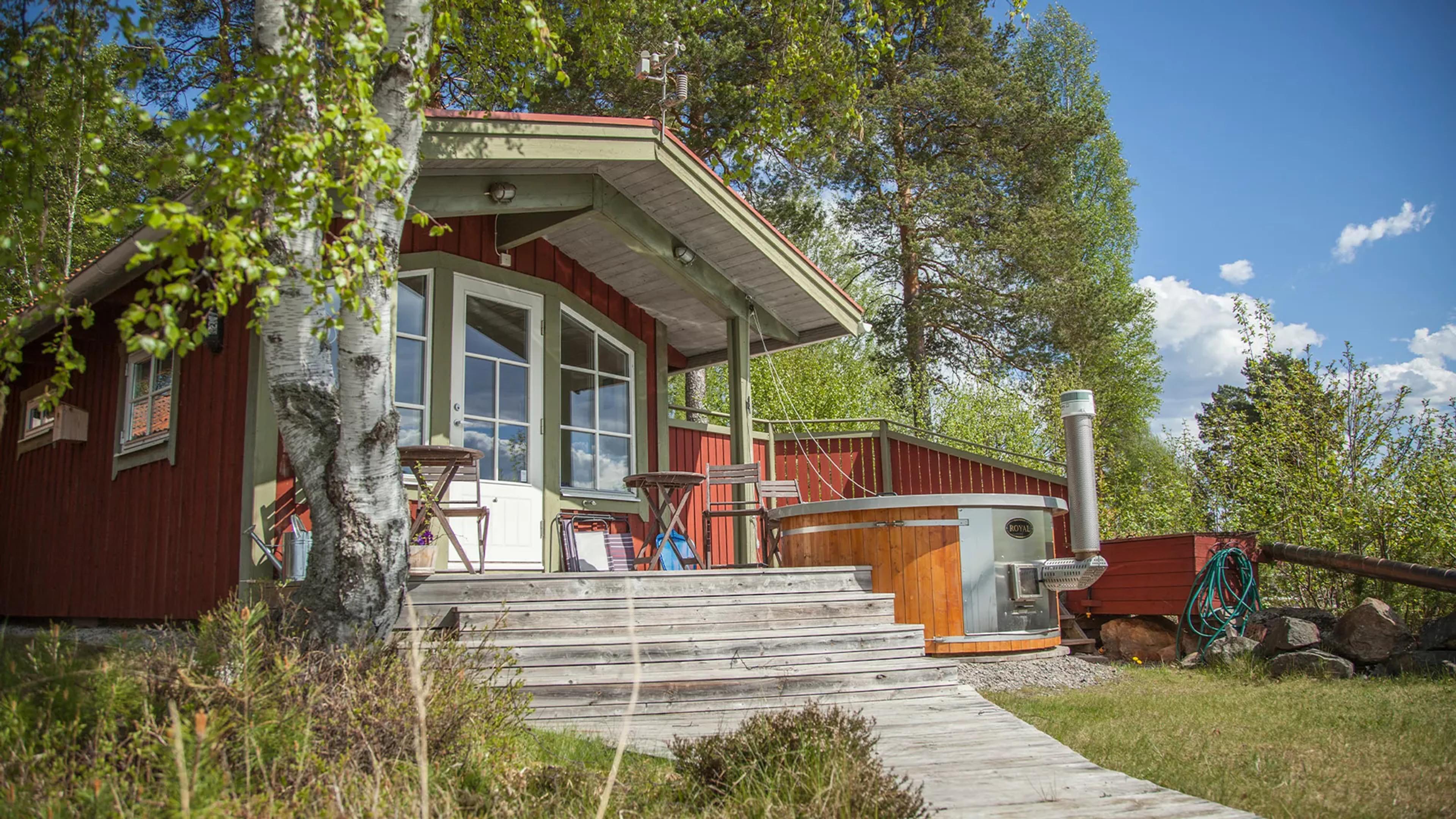 A red cabin with green trim, a wooden deck, and a wood-fired hot tub, surrounded by trees and grass under a blue sky.