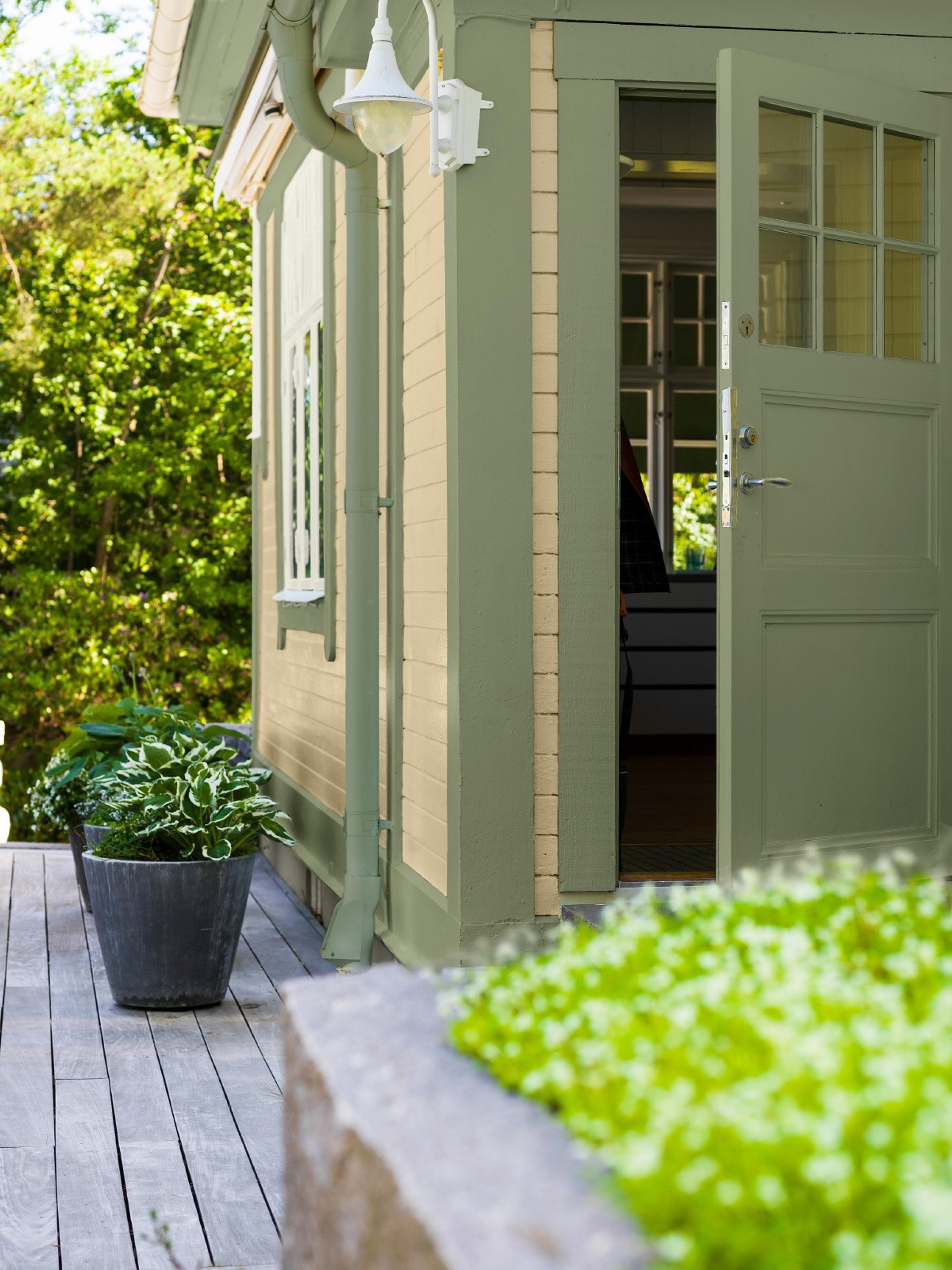 A cream-colored house with green trim and an open green door, leading to a wooden deck with potted plants and a garden.