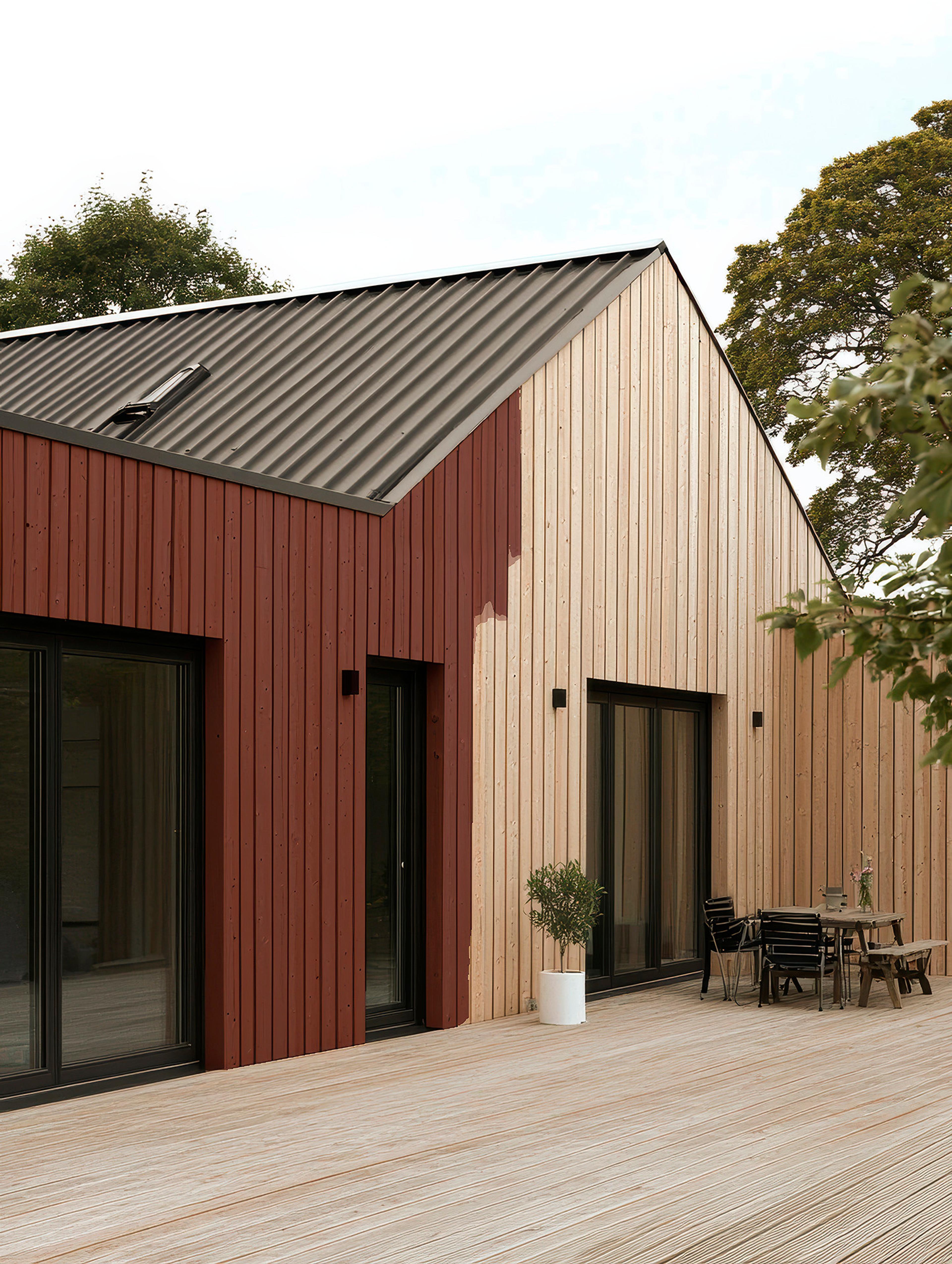 Modern house with a dark metal roof, two-tone wood paneling (reddish-brown and natural wood), and a large wooden deck with outdoor furniture.