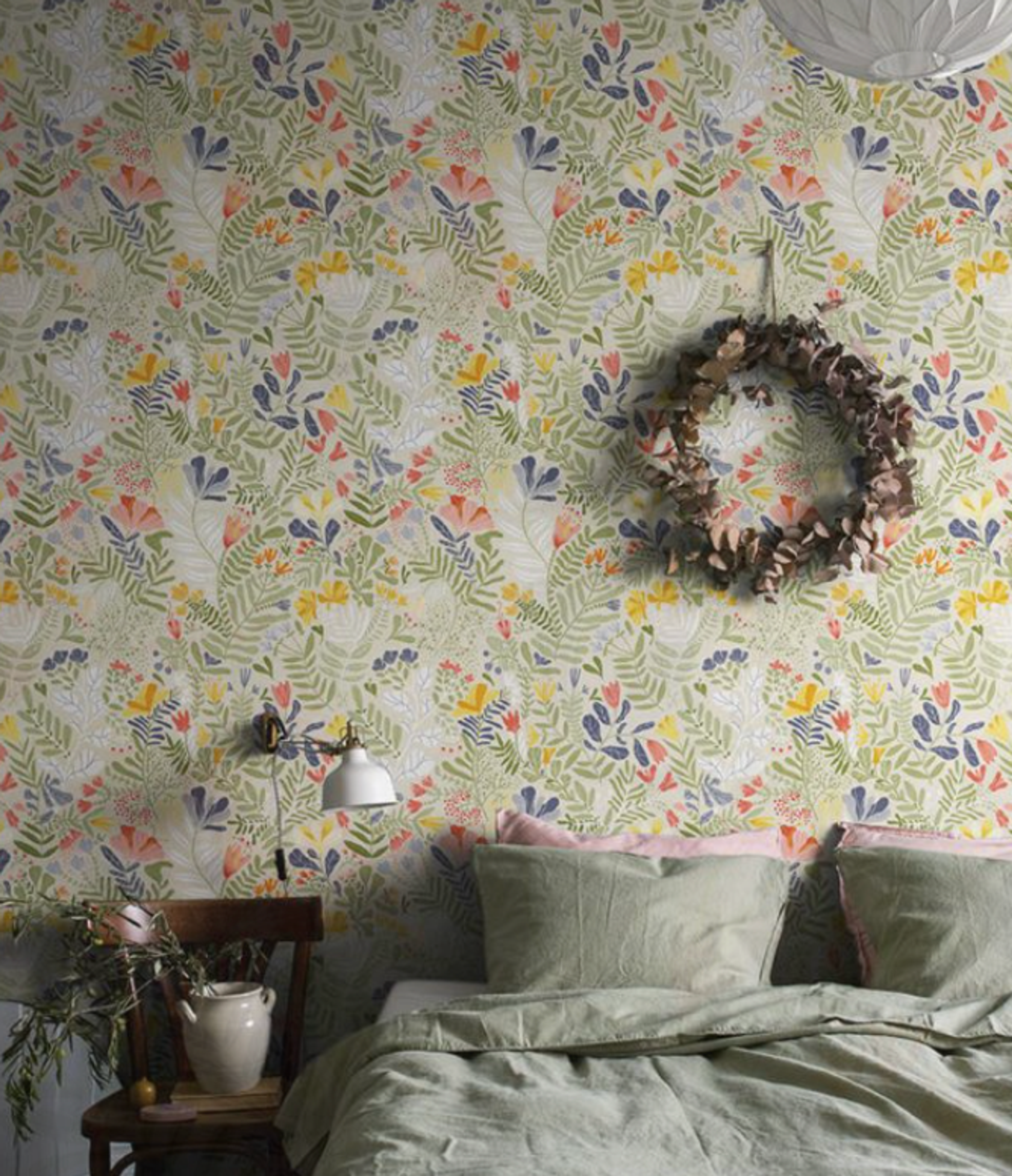 A cozy bedroom featuring colorful floral wallpaper, a bed with sage green bedding, and a dried leaf wreath.