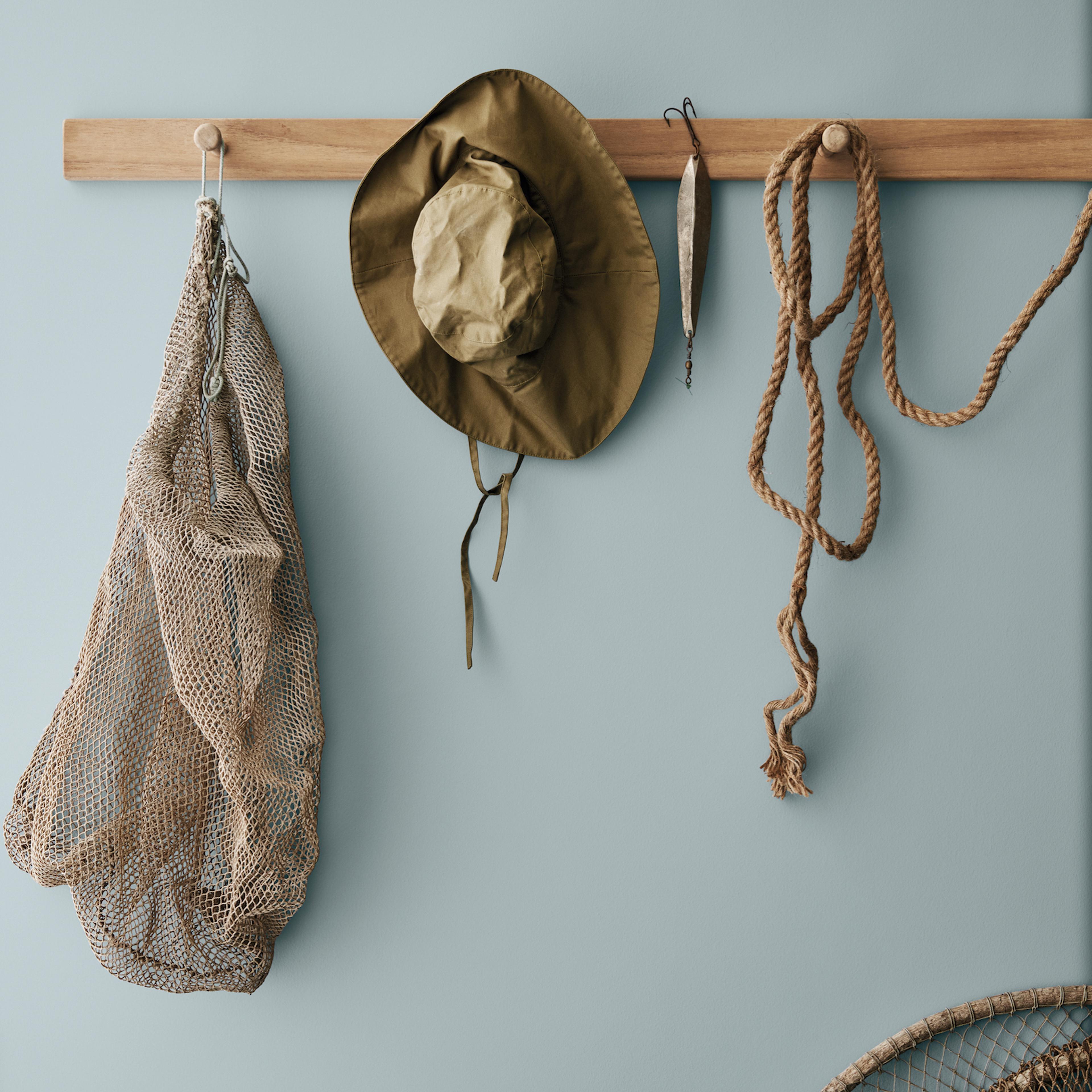 A fishing net, bucket hat, lure, and rope hanging from wooden pegs on a light blue wall.
