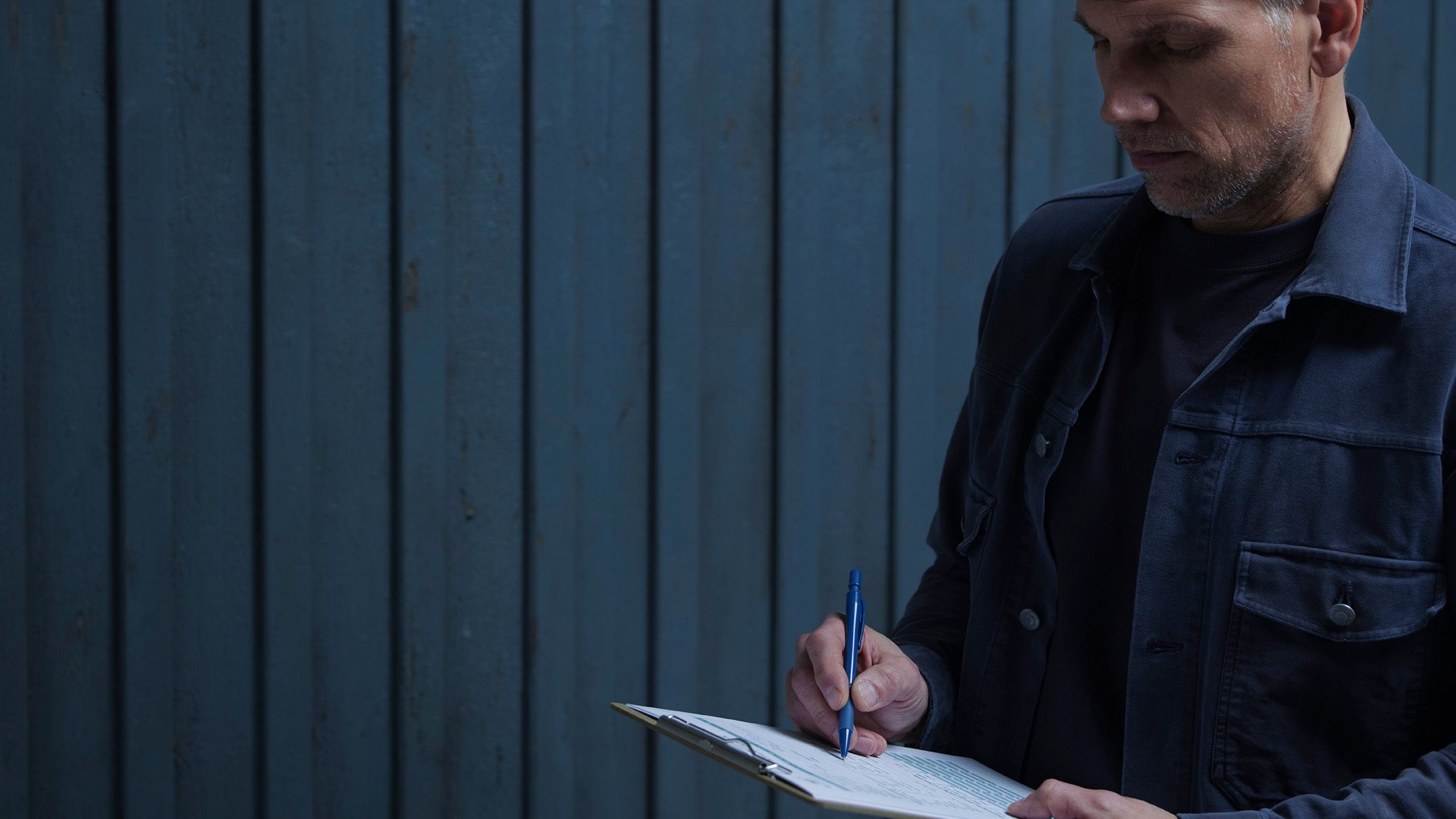 A man in a dark jacket writes on a clipboard with a blue pen against a dark blue paneled wall.