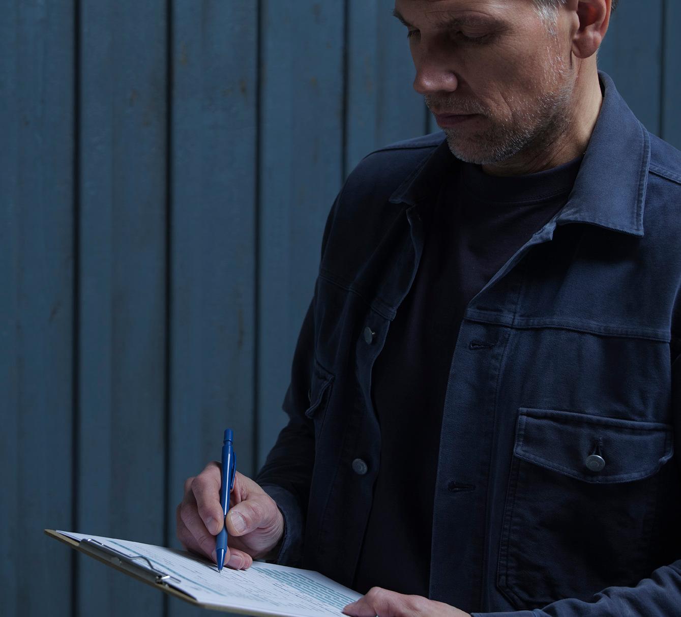A man in a dark jacket writes on a clipboard with a blue pen against a dark blue paneled wall.
