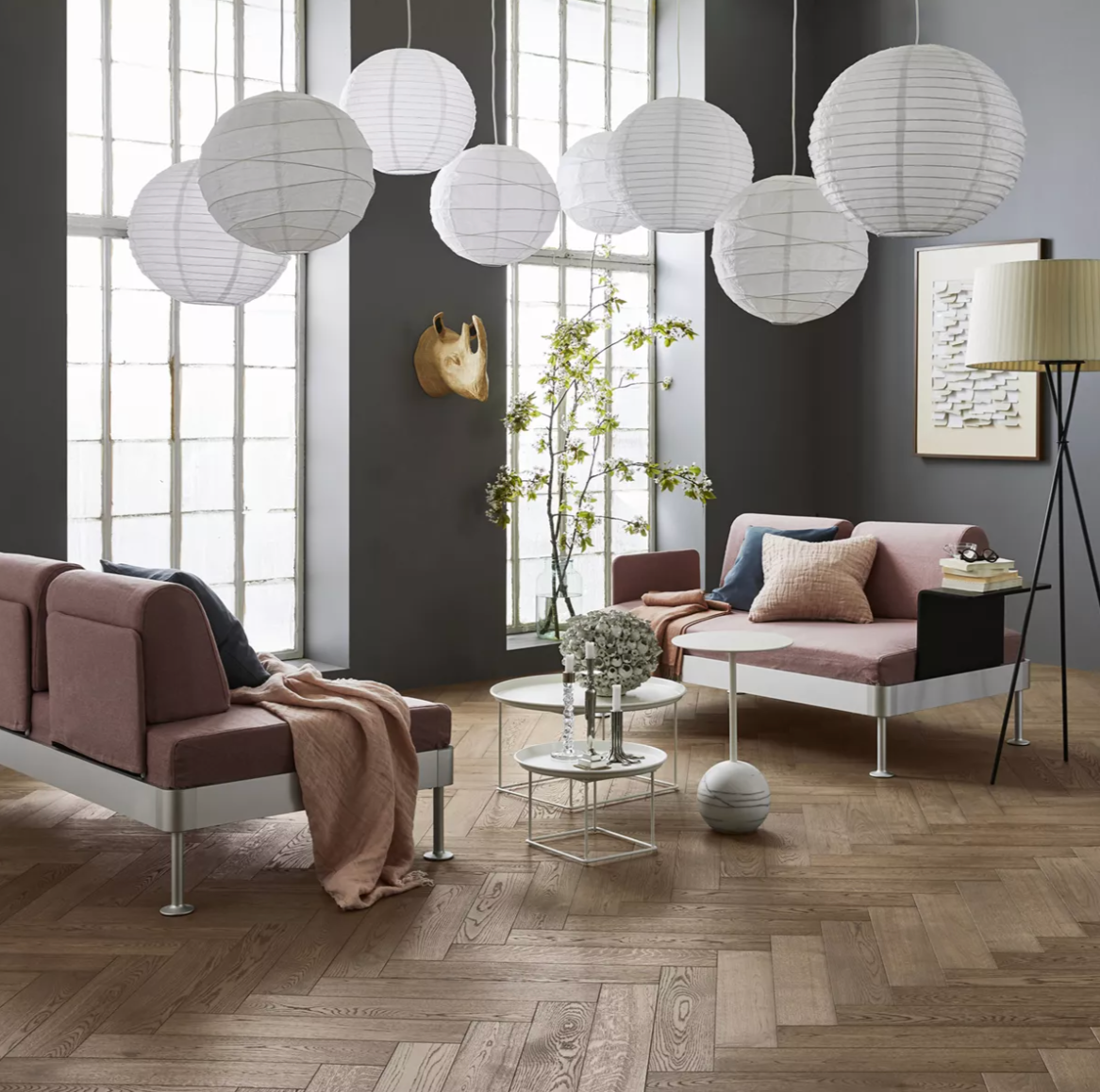 A modern living room with dark grey walls, pink daybeds, white paper lanterns, and a herringbone wood floor.