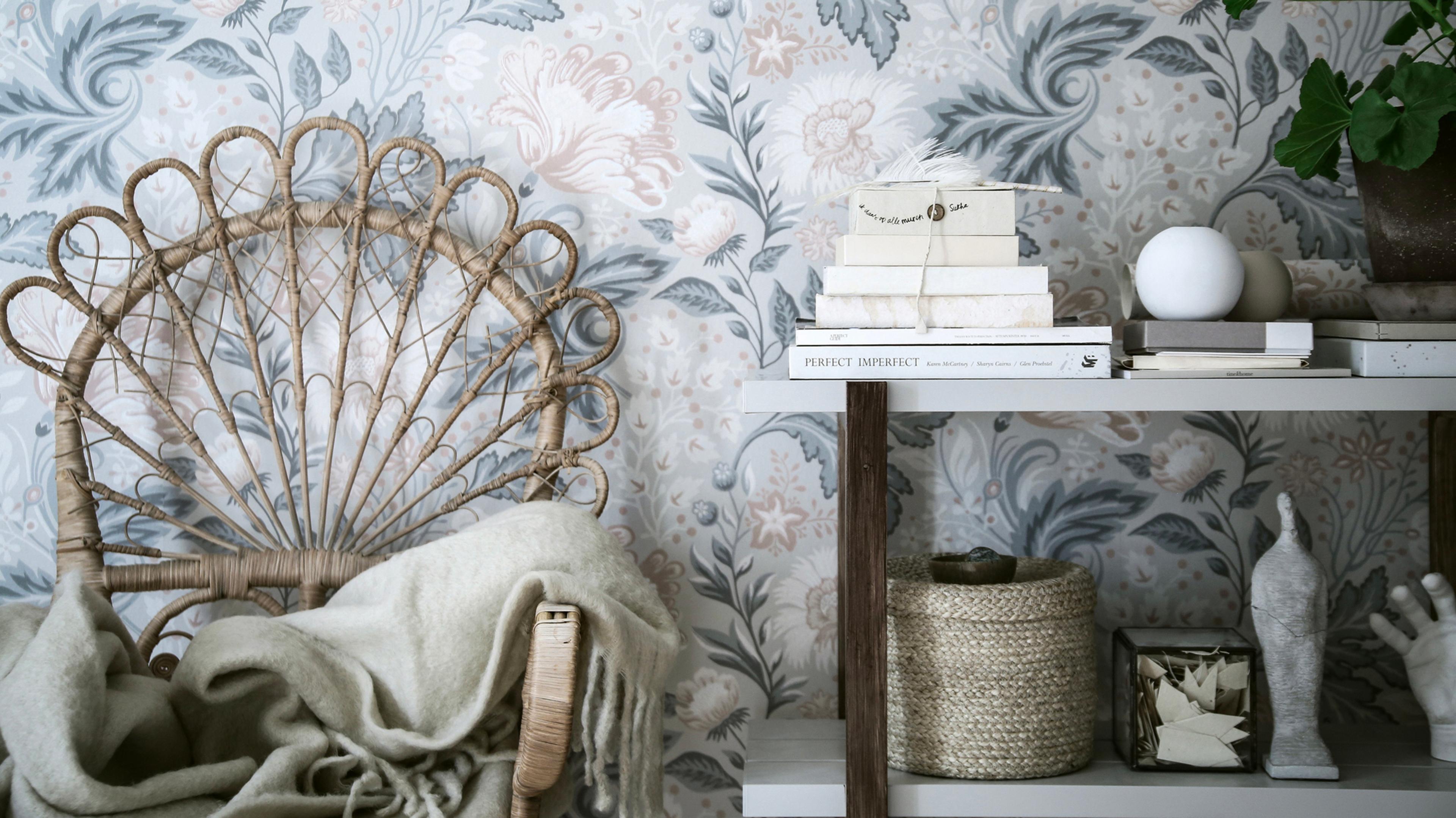 A rattan chair with a blanket and a shelf of decorative items against blue and pink floral wallpaper.
