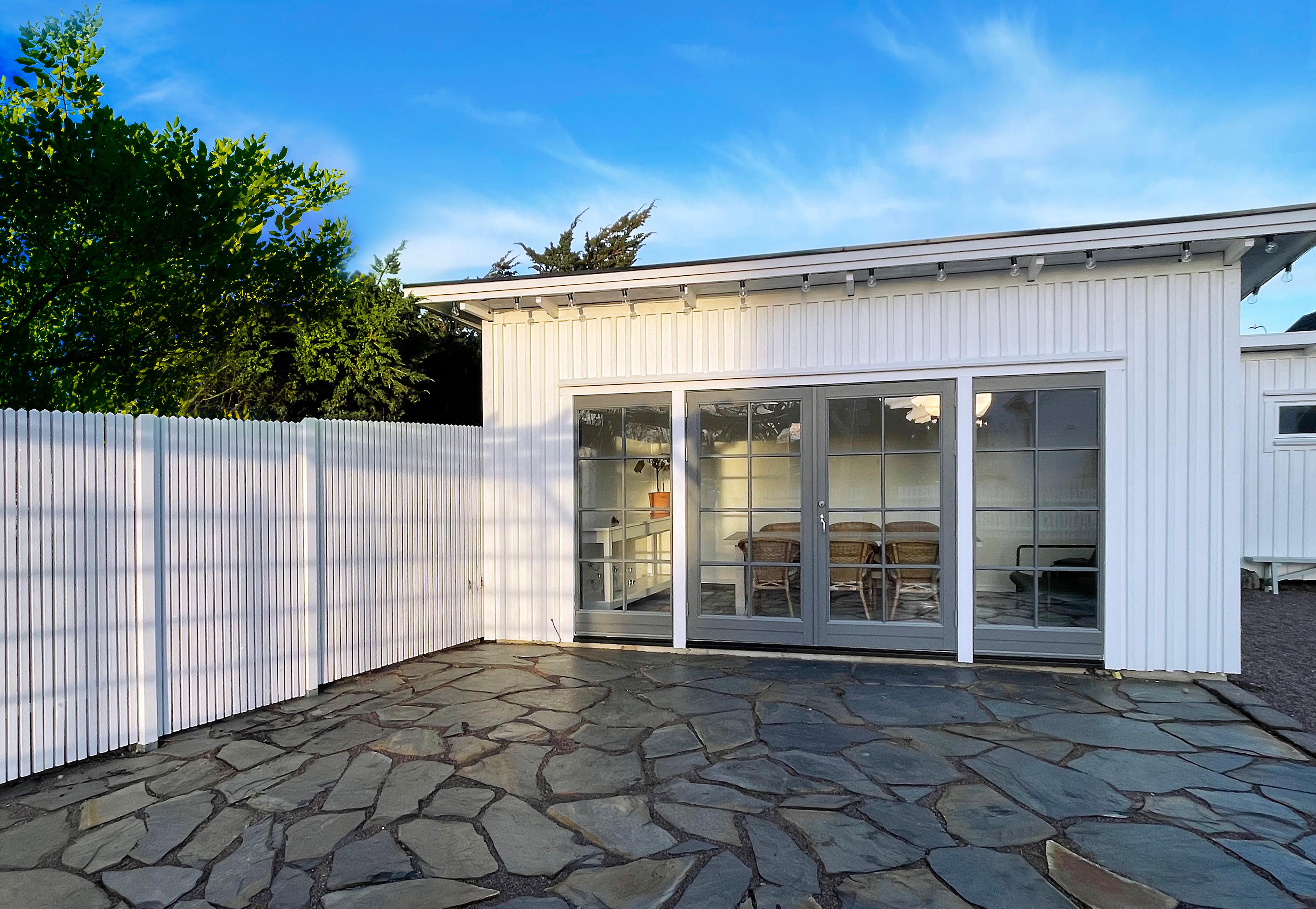 A white building with large glass doors overlooks a patio paved with irregular dark stones, next to a white fence under a blue sky.