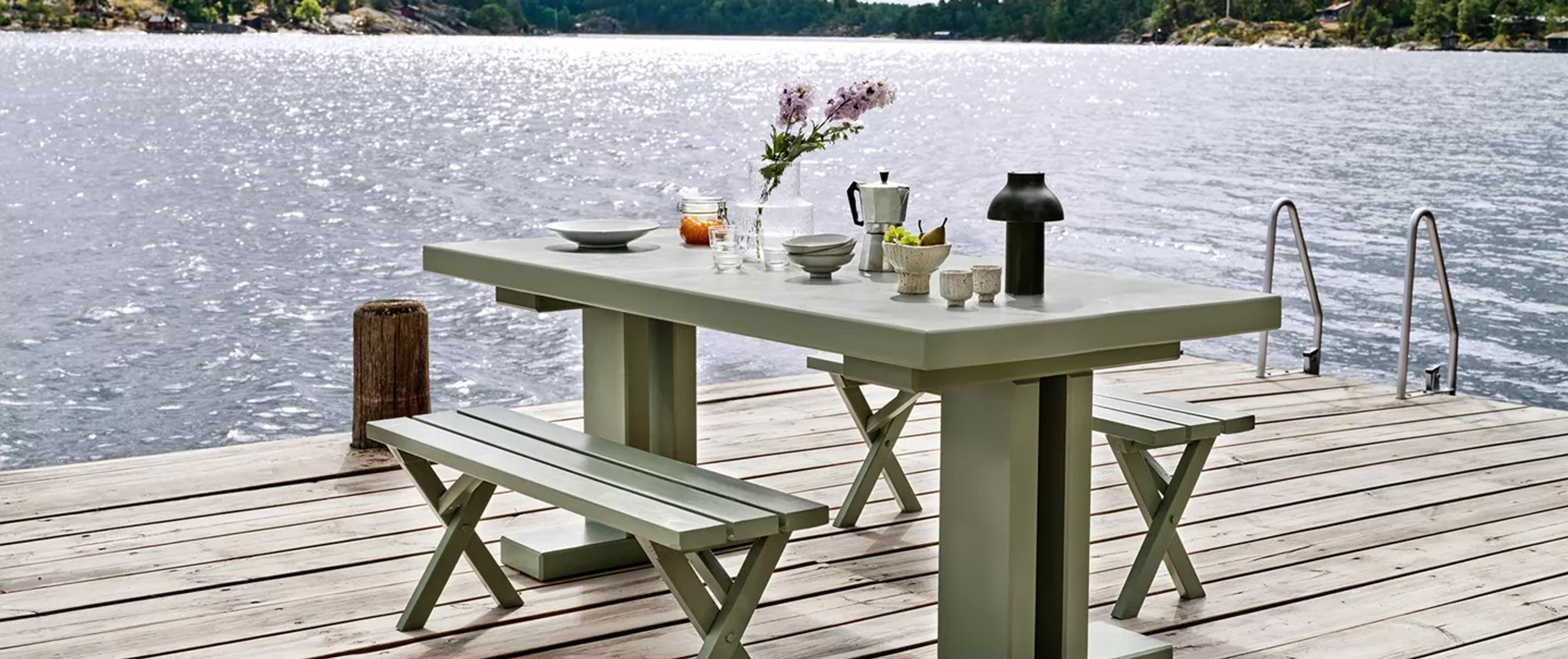 Light green outdoor dining set on a wooden dock by a shimmering lake, adorned with dishes and flowers.