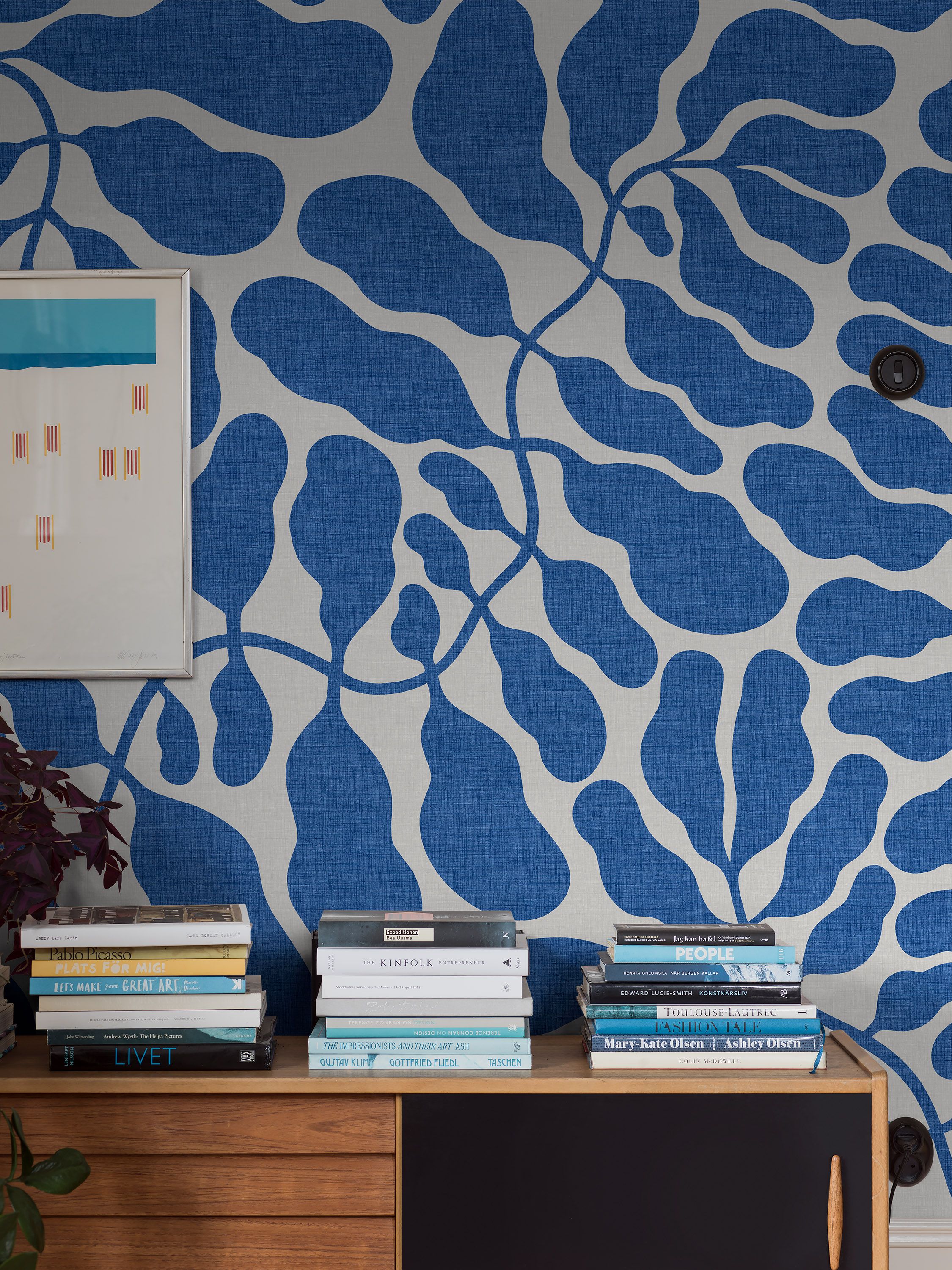 A wooden cabinet topped with several stacks of books sits against a wall with blue and off-white abstract organic pattern wallpaper, with a framed artwork on the left.