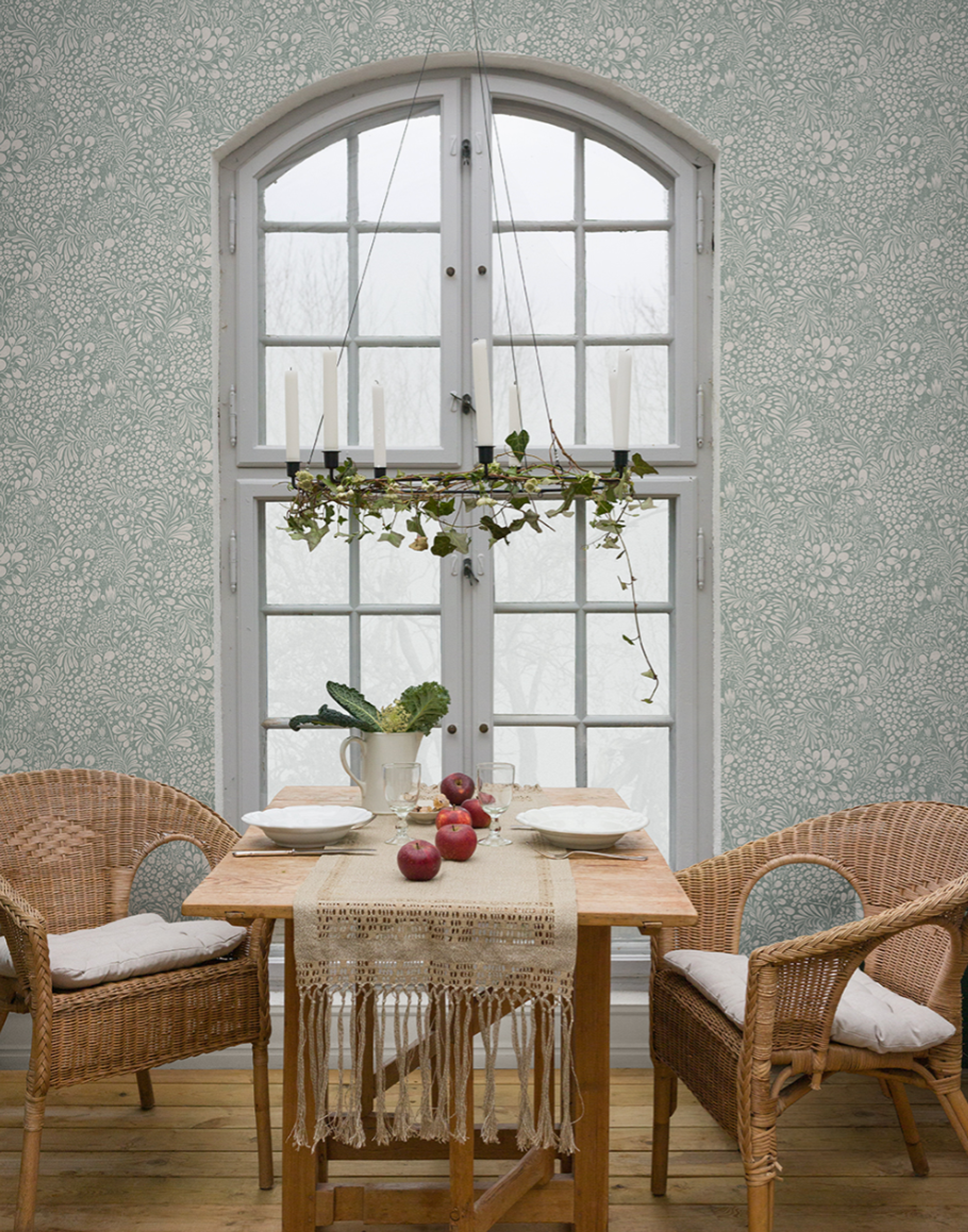 A dining room with light green floral wallpaper, a wooden table set for two with wicker chairs, and an arched window featuring a hanging candle holder decorated with ivy.