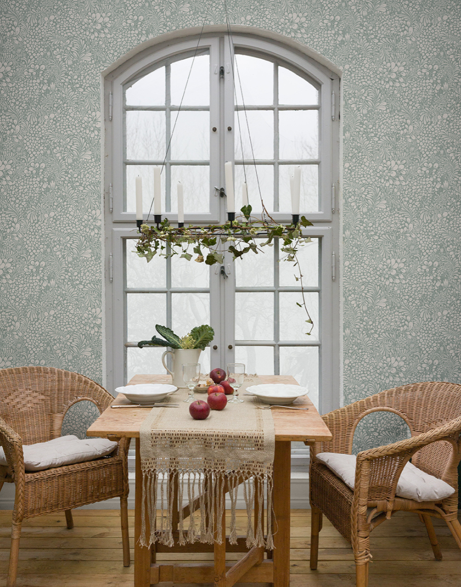A dining room with light green floral wallpaper, a wooden table set for two with wicker chairs, and an arched window featuring a hanging candle holder decorated with ivy.
