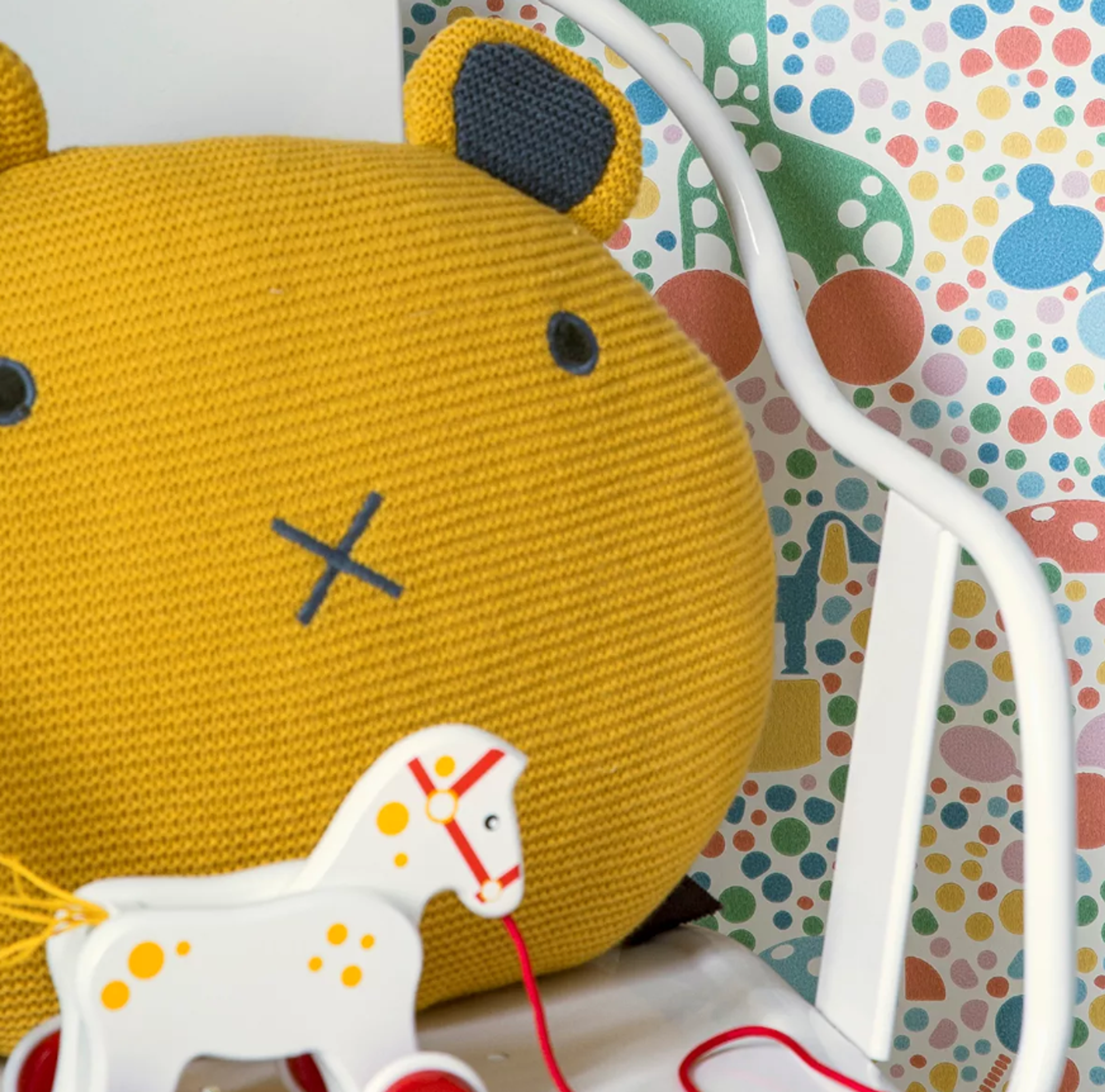 A yellow knitted bear cushion, a white wooden pull-along horse toy, and colorful wallpaper with polka dots and playful illustrations, next to a white crib rail.