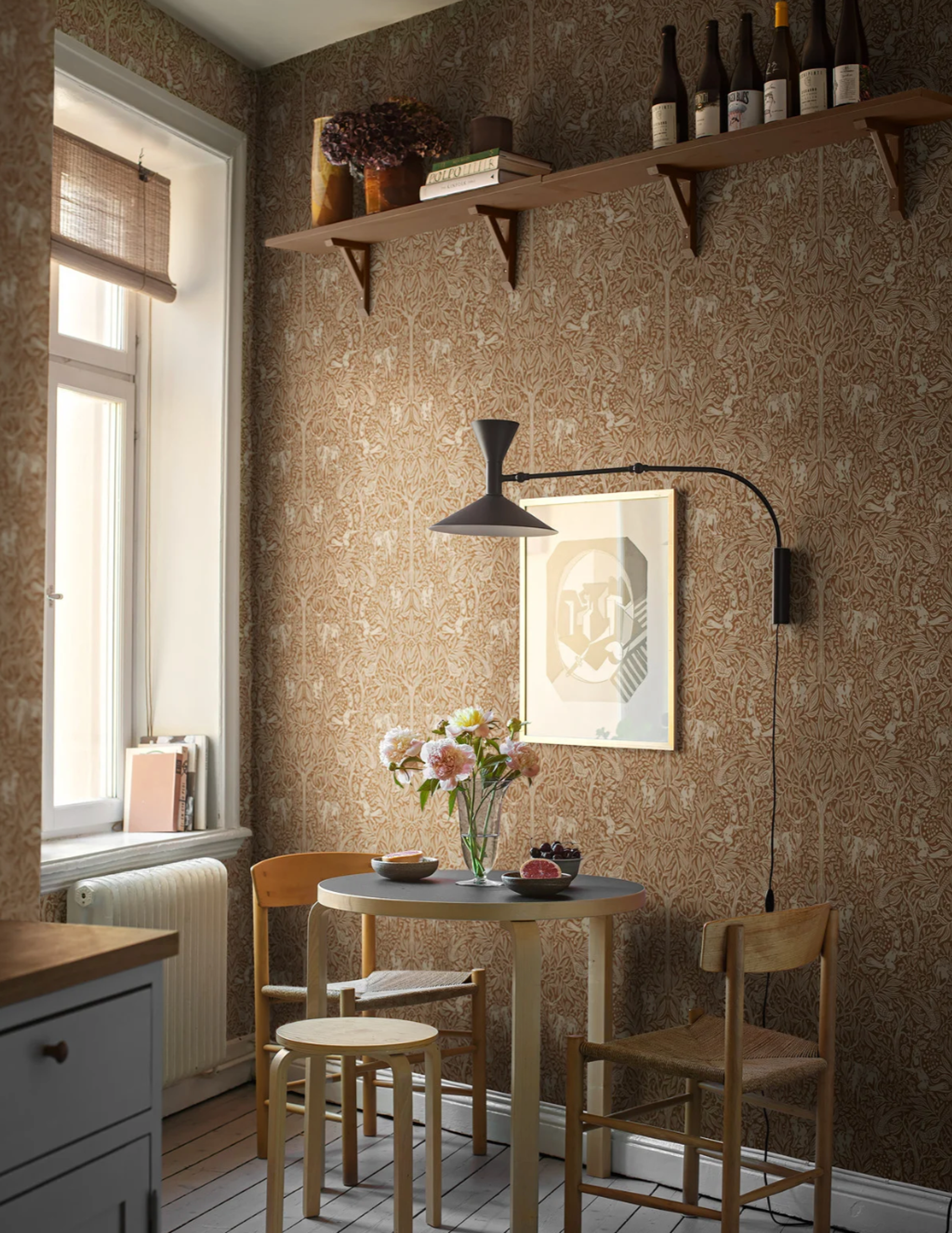 A cozy dining nook featuring light brown tree-patterned wallpaper, a small round table with flowers, and wall shelves.