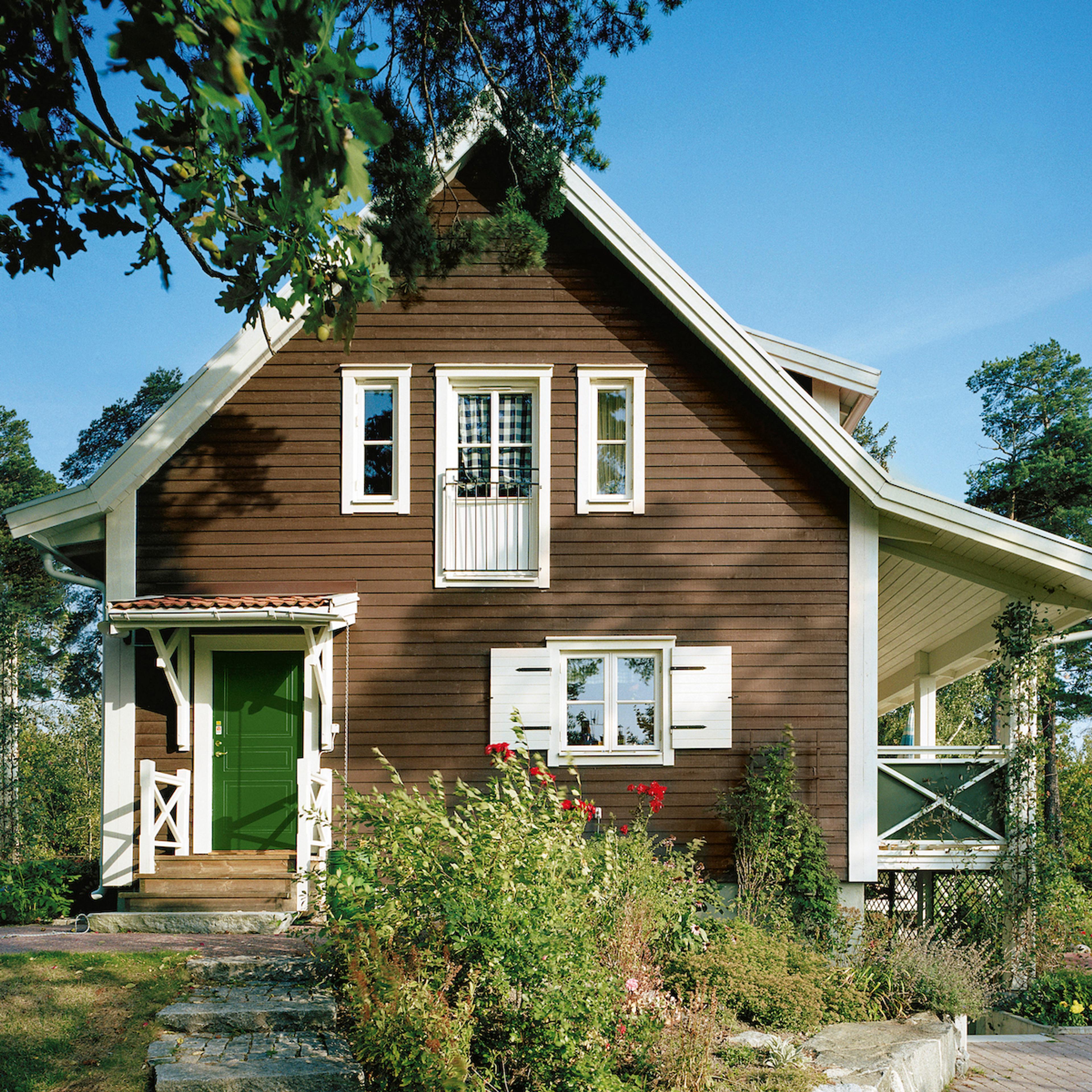 A dark brown two-story wooden house with a green front door, white window trim, white shutters, and a small balcony, set in a lush garden under a clear blue sky.