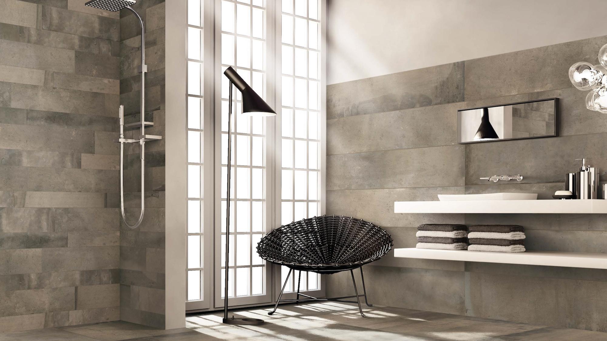 A modern bathroom with large grey tiles, a shower, a white floating vanity, and a black wire chair next to a large window.
