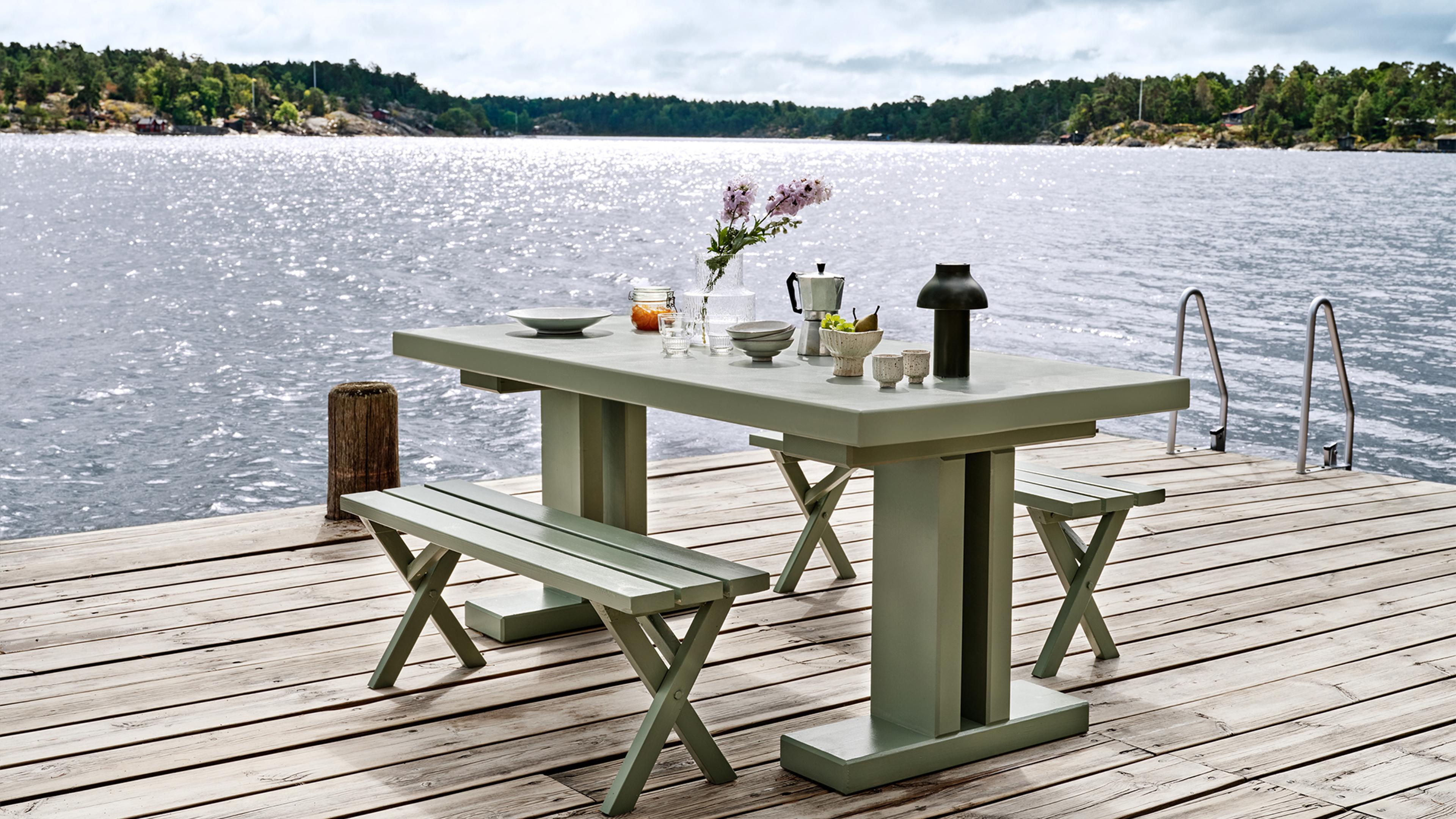 A light green outdoor dining set on a wooden dock by a lake, with the table arranged with dishes, flowers, and a lamp.