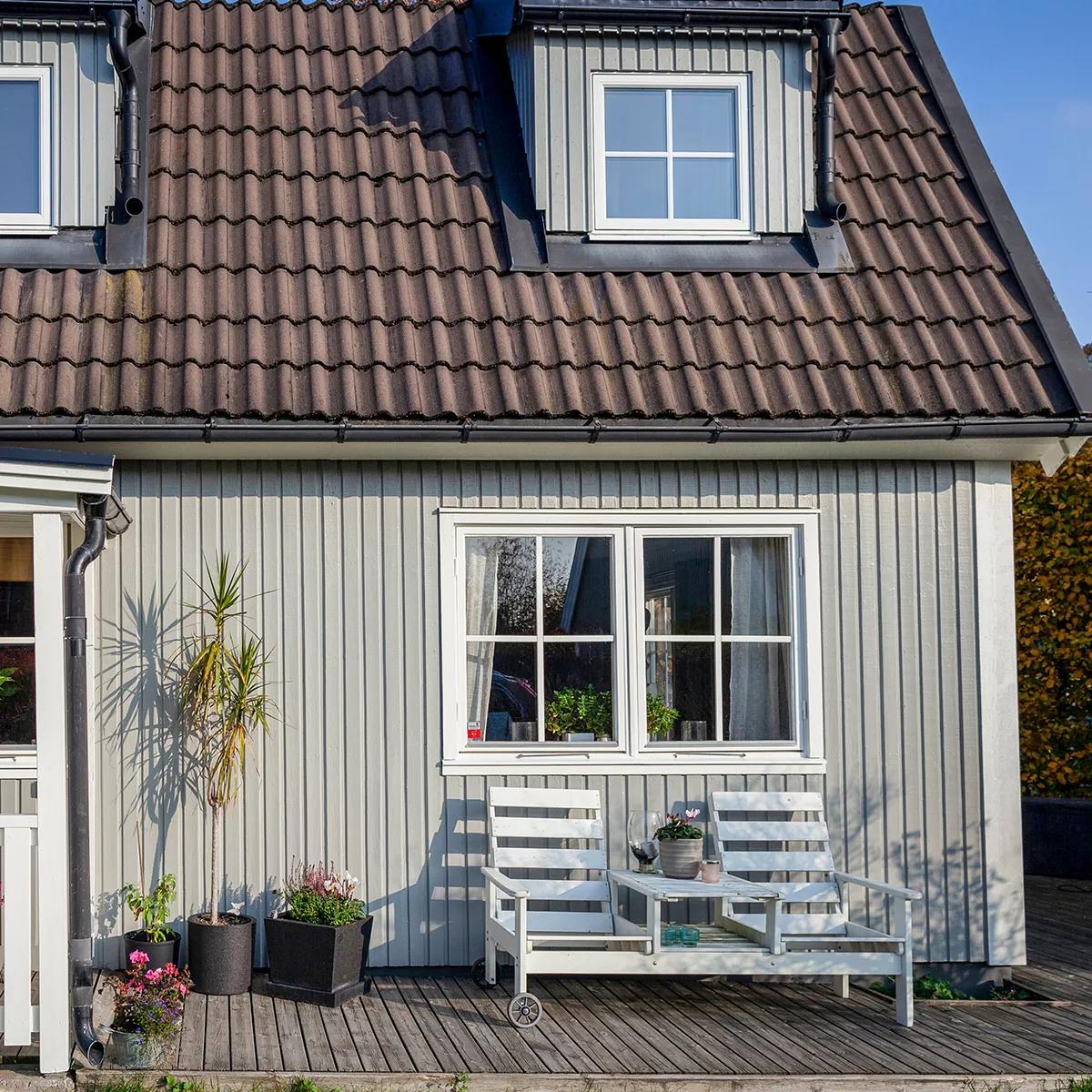 Light gray house with a brown tiled roof, dormer windows, and a wooden deck featuring a white patio set and potted plants.
