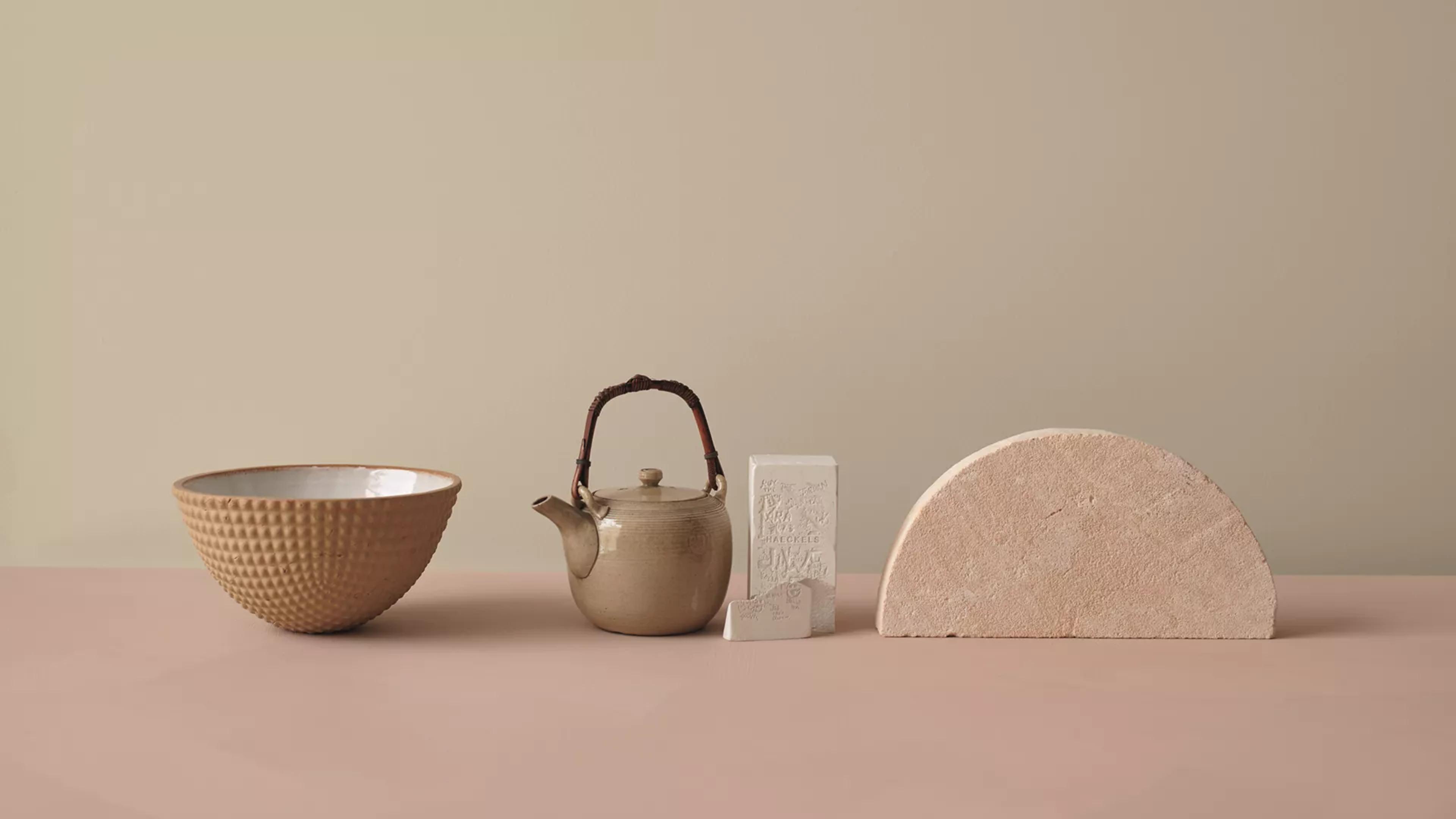 A still life featuring a textured terracotta bowl, a beige teapot, two carved white blocks, and a large pink semi-circle stone on a pink surface.