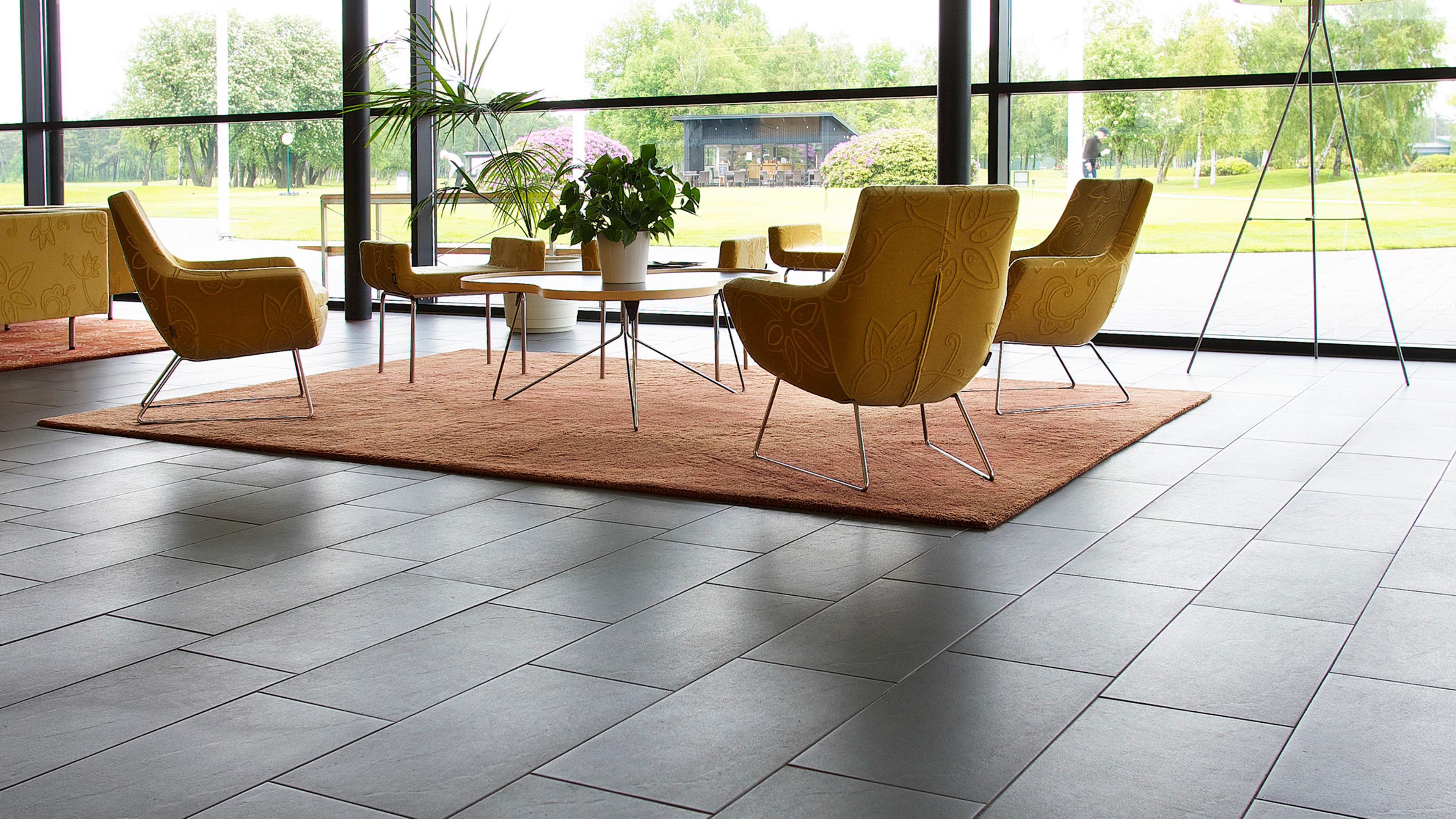 A modern lounge area with dark grey tiled floors, mustard yellow armchairs, a rust-colored rug, and large windows overlooking a green landscape.