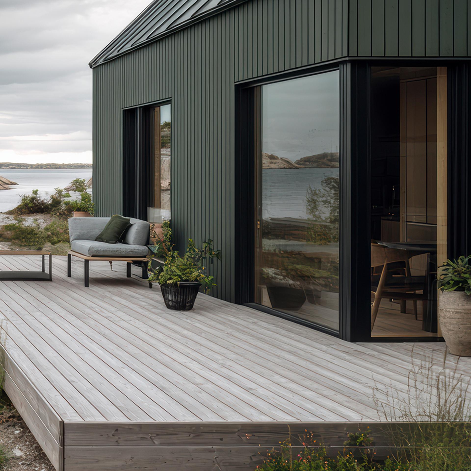 Modern dark green house with a wooden deck and outdoor seating overlooking a rocky bay.