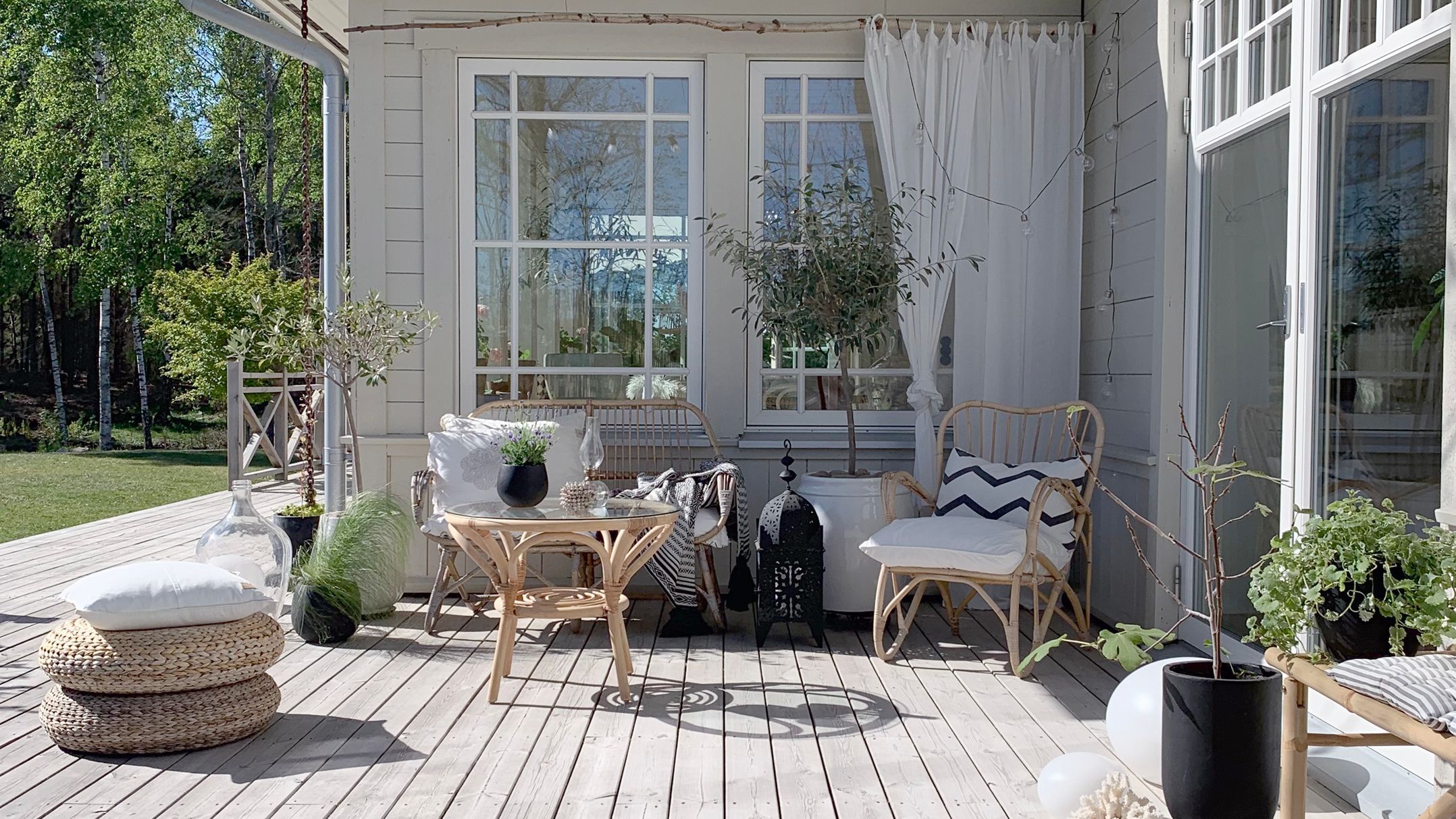 A cozy outdoor veranda with wicker furniture, potted plants, and large white-framed windows overlooking a green lawn.