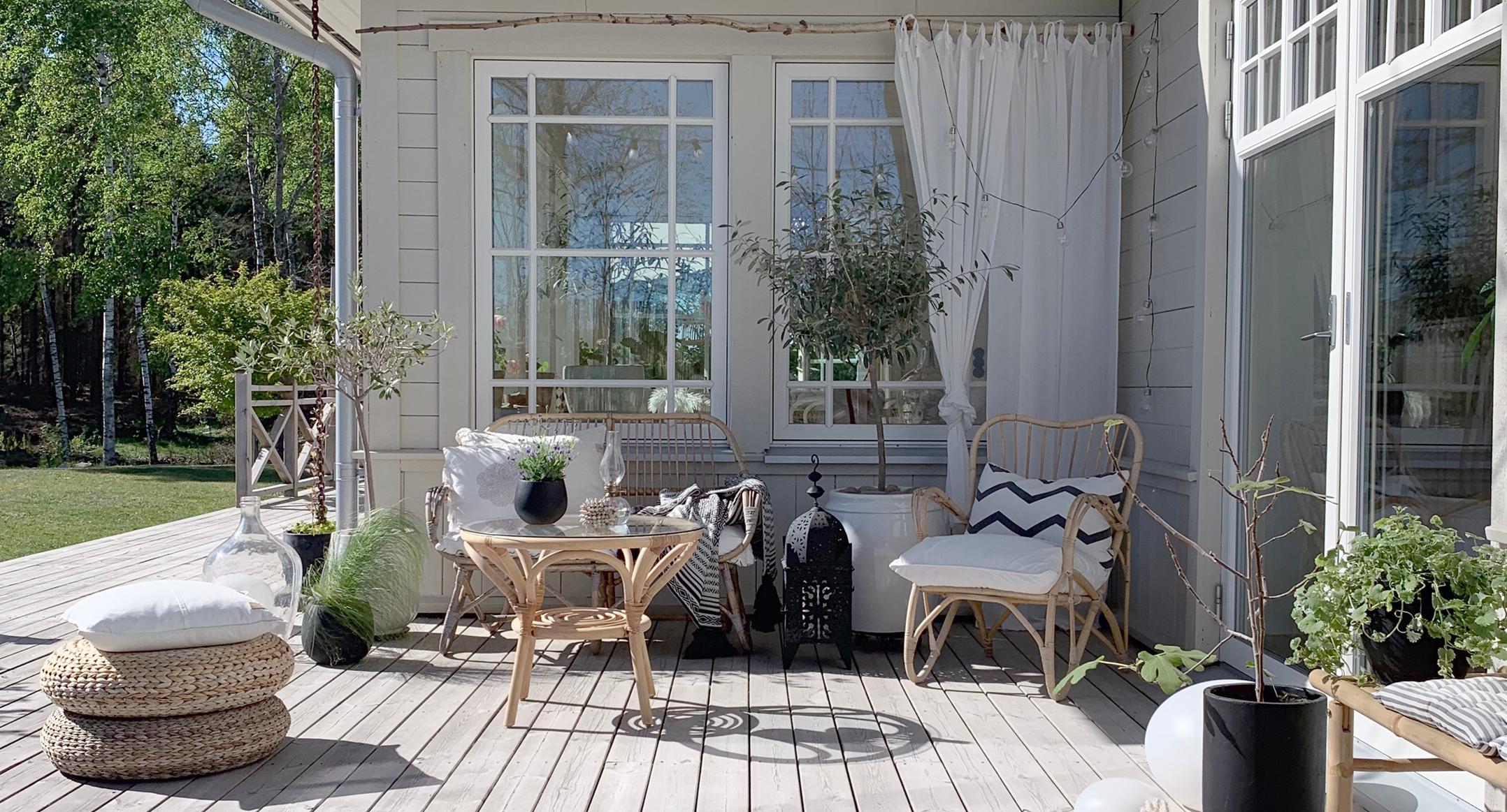 A cozy outdoor veranda with wicker furniture, potted plants, and large white-framed windows overlooking a green lawn.