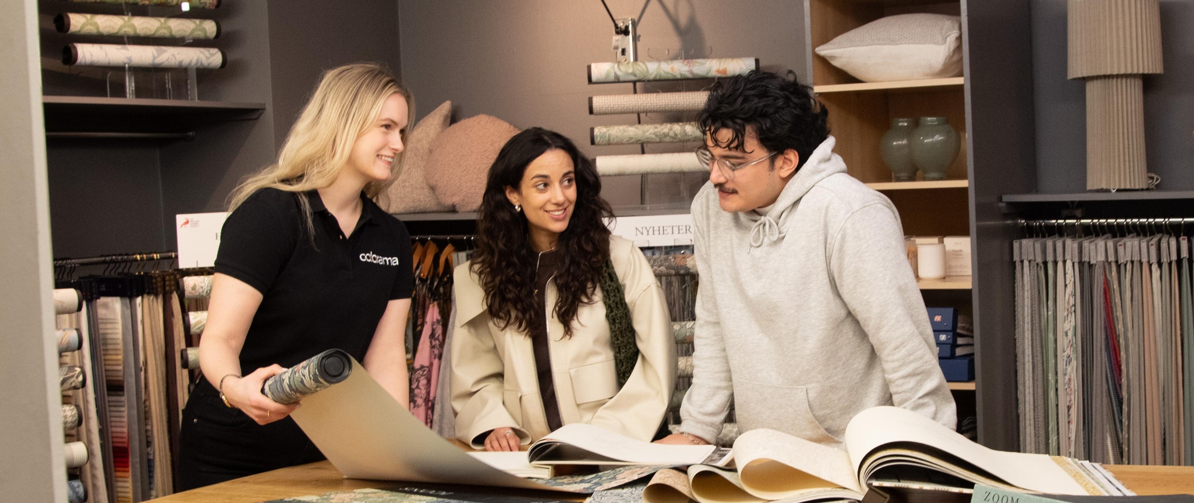 An employee helps two customers look at wallpaper and fabric samples in a home decor store.