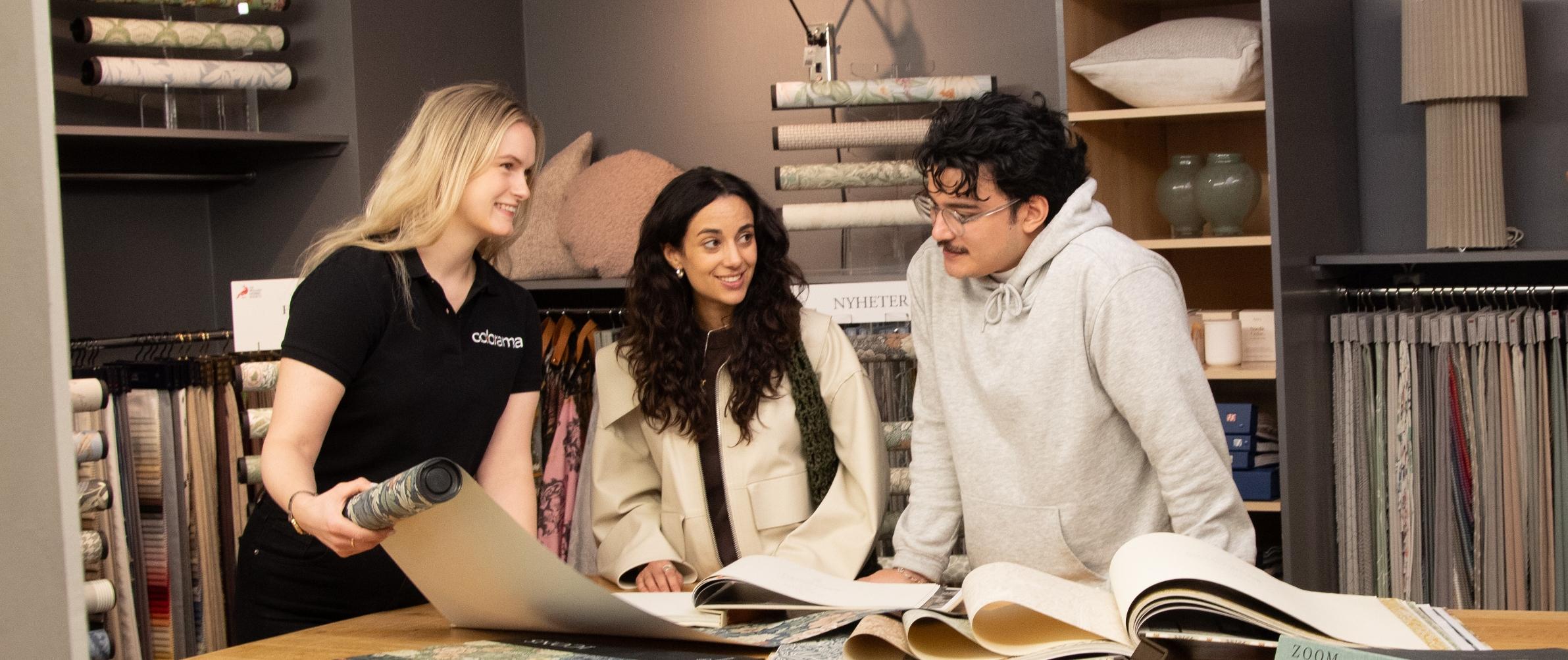 An employee helps two customers look at wallpaper and fabric samples in a home decor store.