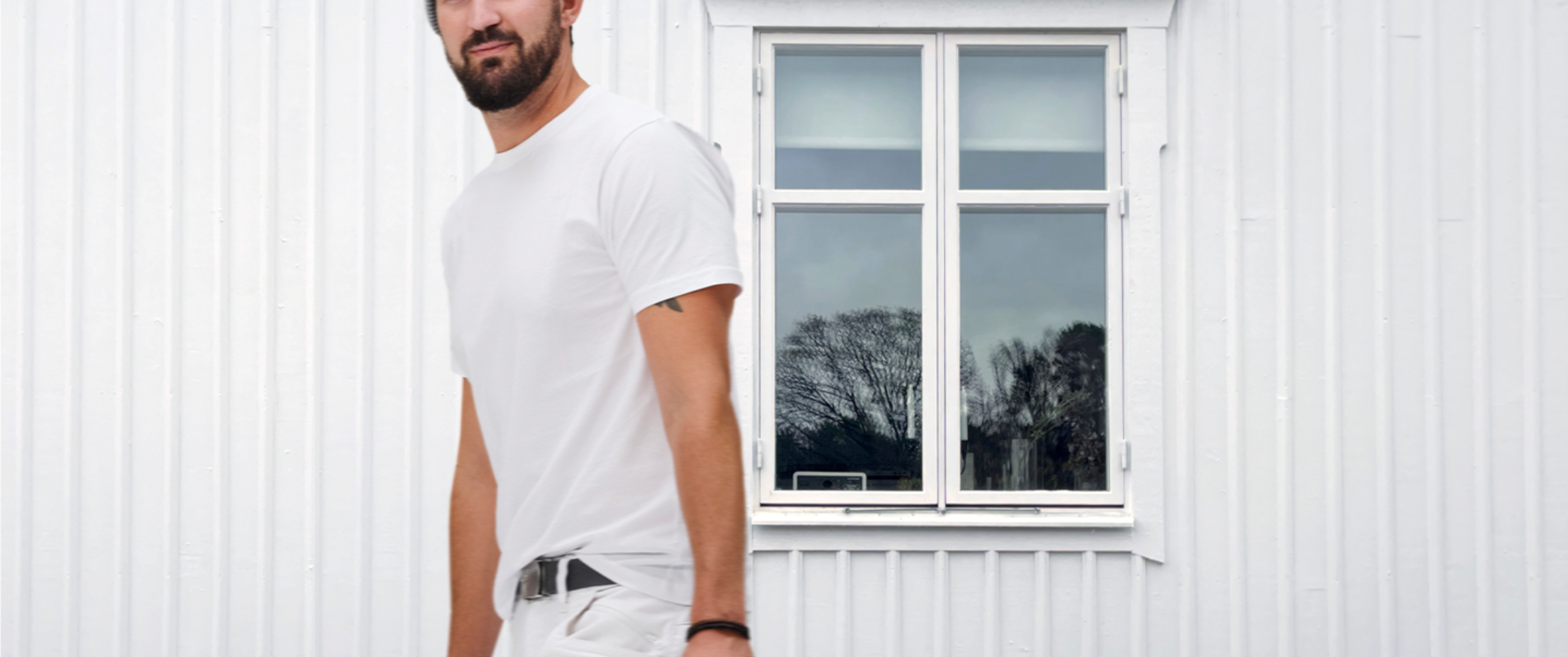 White-clad man with a beard stands next to a white building with a window reflecting trees.