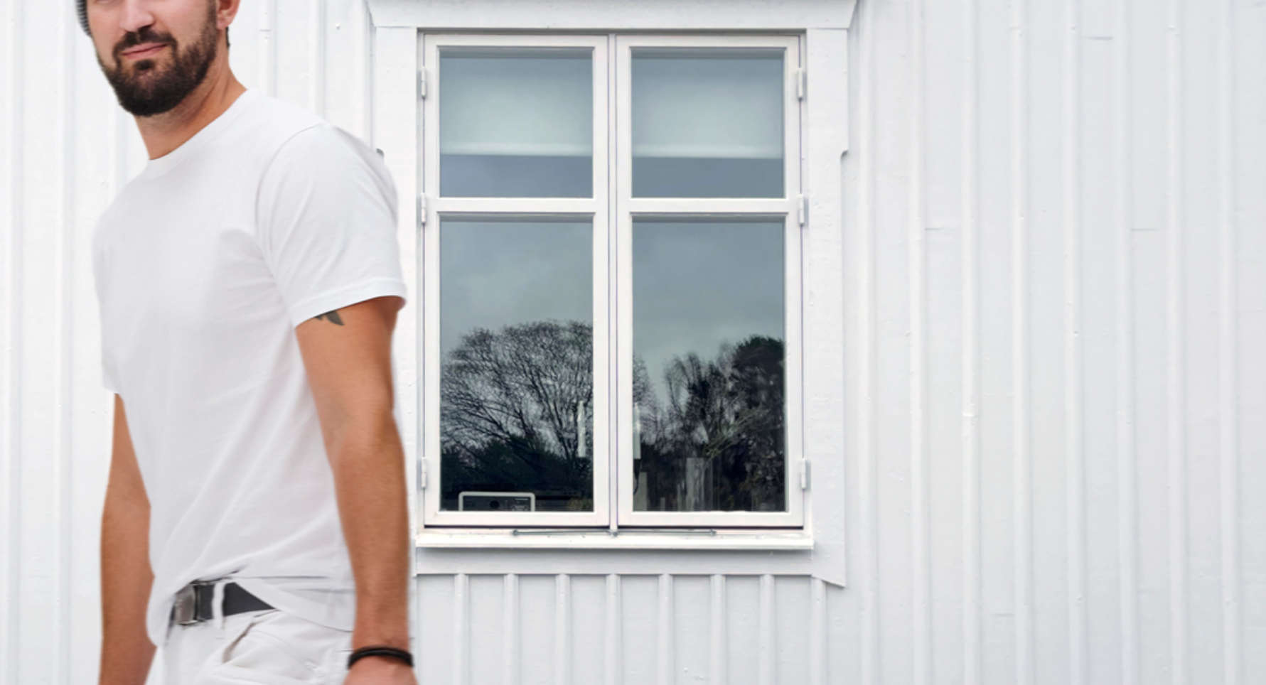 White-clad man with a beard stands next to a white building with a window reflecting trees.