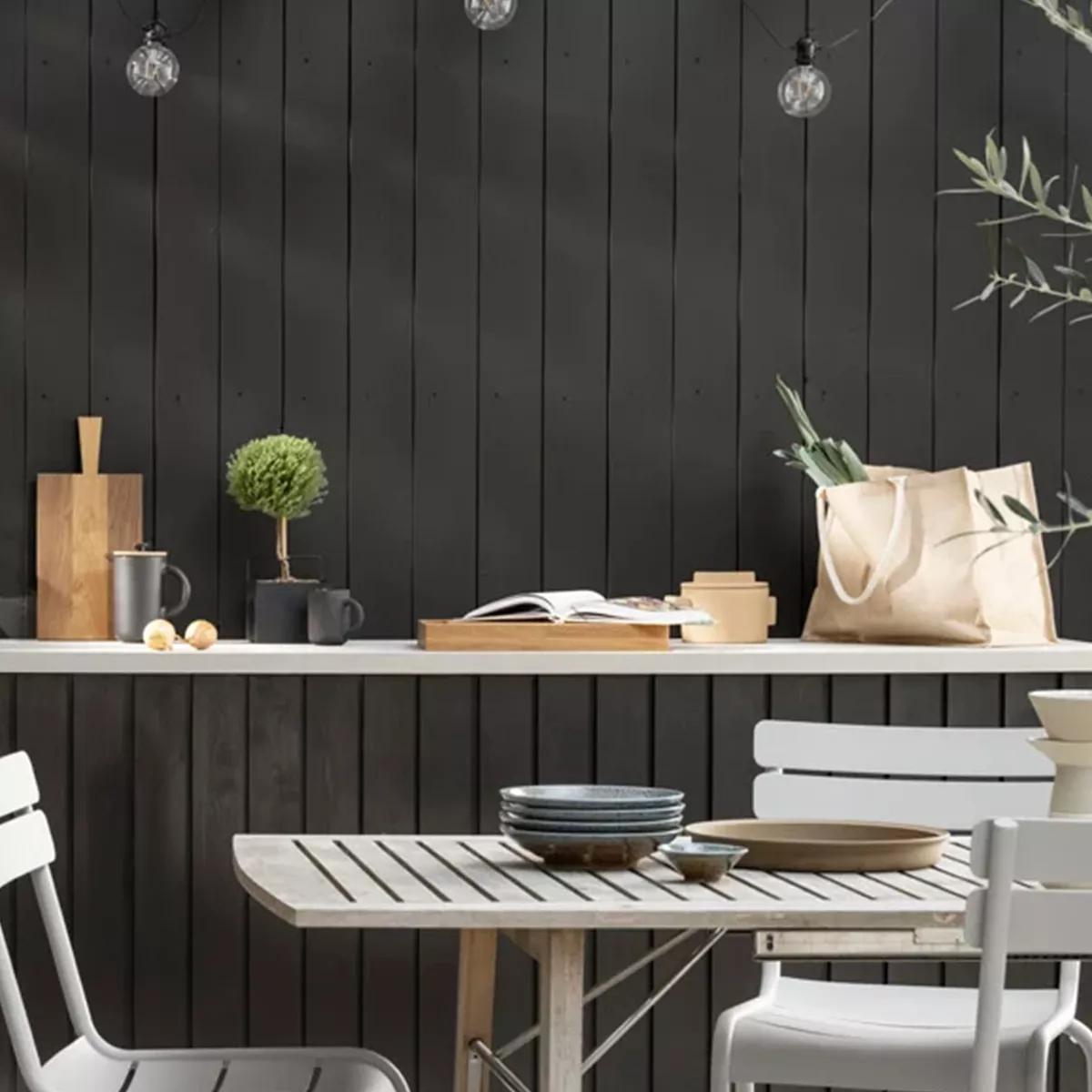 A modern outdoor patio features a dark wood paneled wall, a white shelf with various items, and a slatted dining table with plates and white chairs.