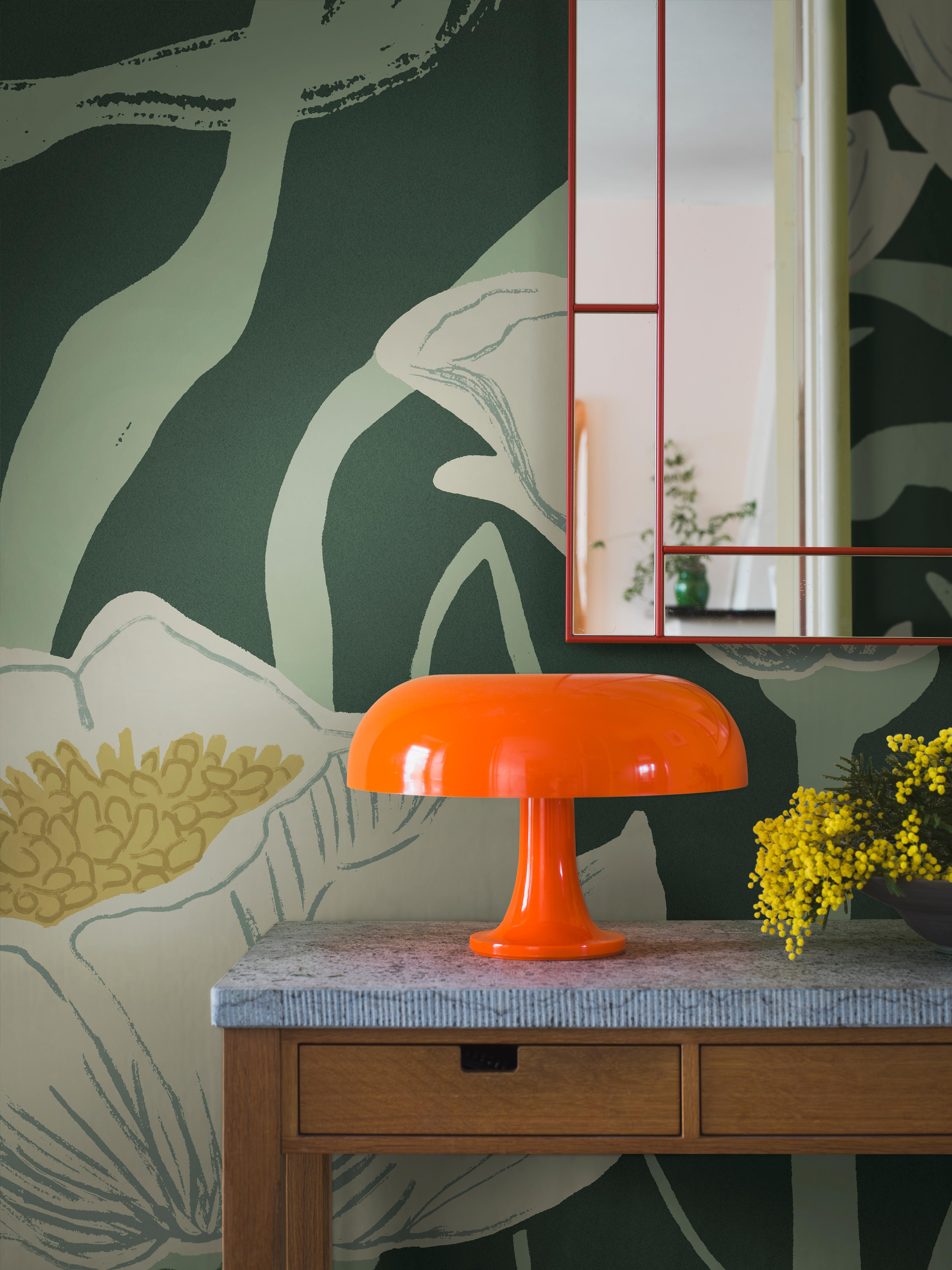 A vibrant orange mushroom lamp and yellow mimosa flowers sit on a wooden console table with a grey top, against a dark green floral wallpaper with a red-framed mirror above.
