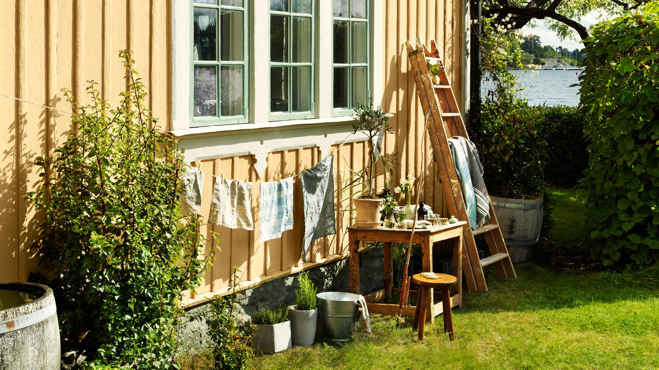 A yellow wooden cottage with clothes drying outside, a garden, and a lake in the background.