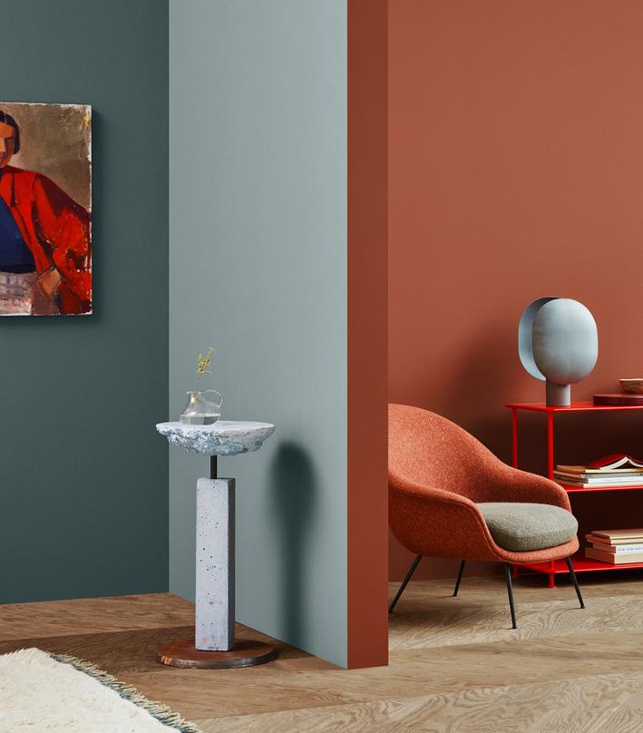 A modern interior with color-blocked walls in teal, light blue, and terracotta, featuring an orange armchair, a concrete pedestal table, and a red shelving unit.