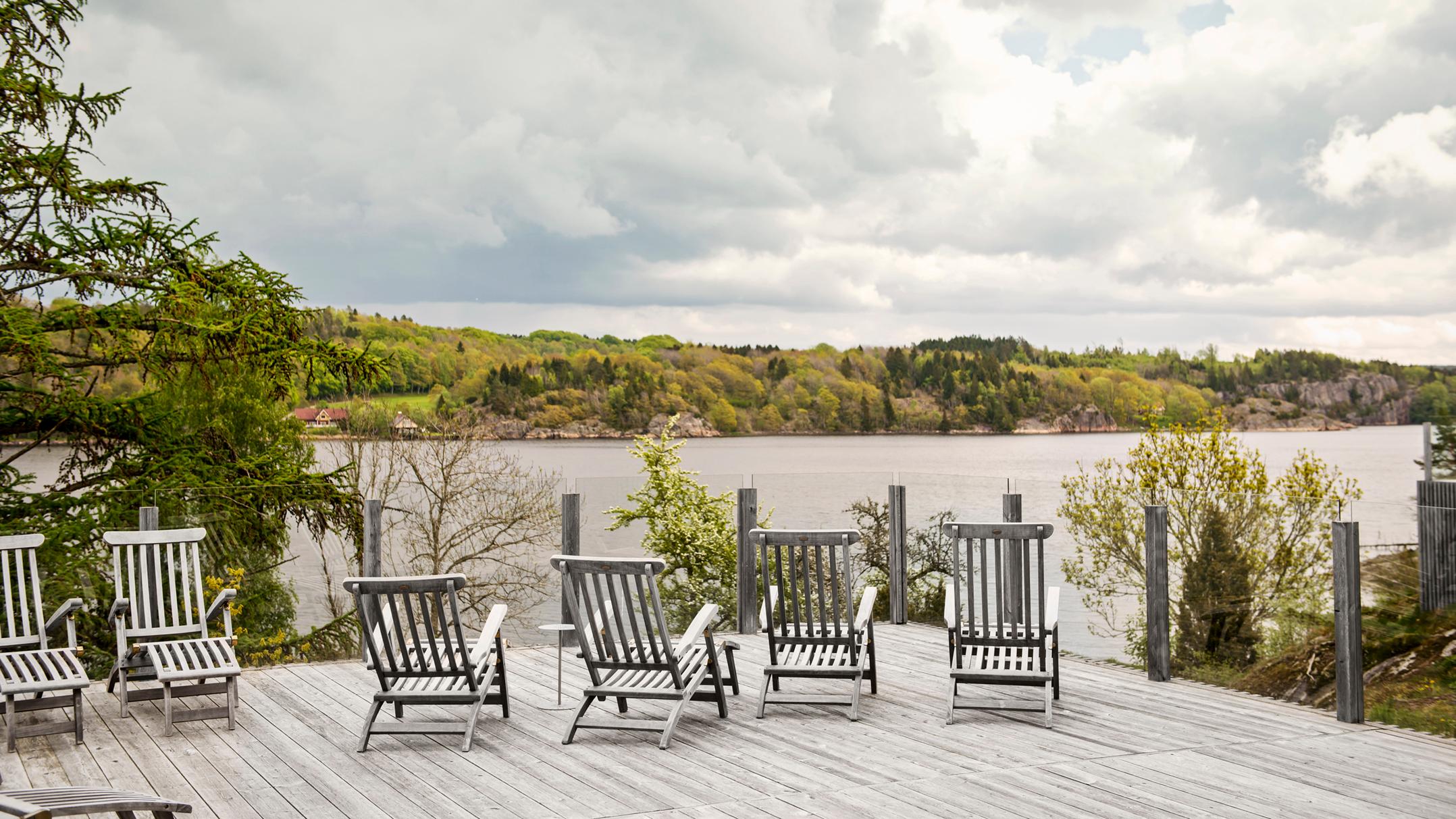 A wooden deck with lounge chairs overlooks a lake surrounded by green trees under a cloudy sky.