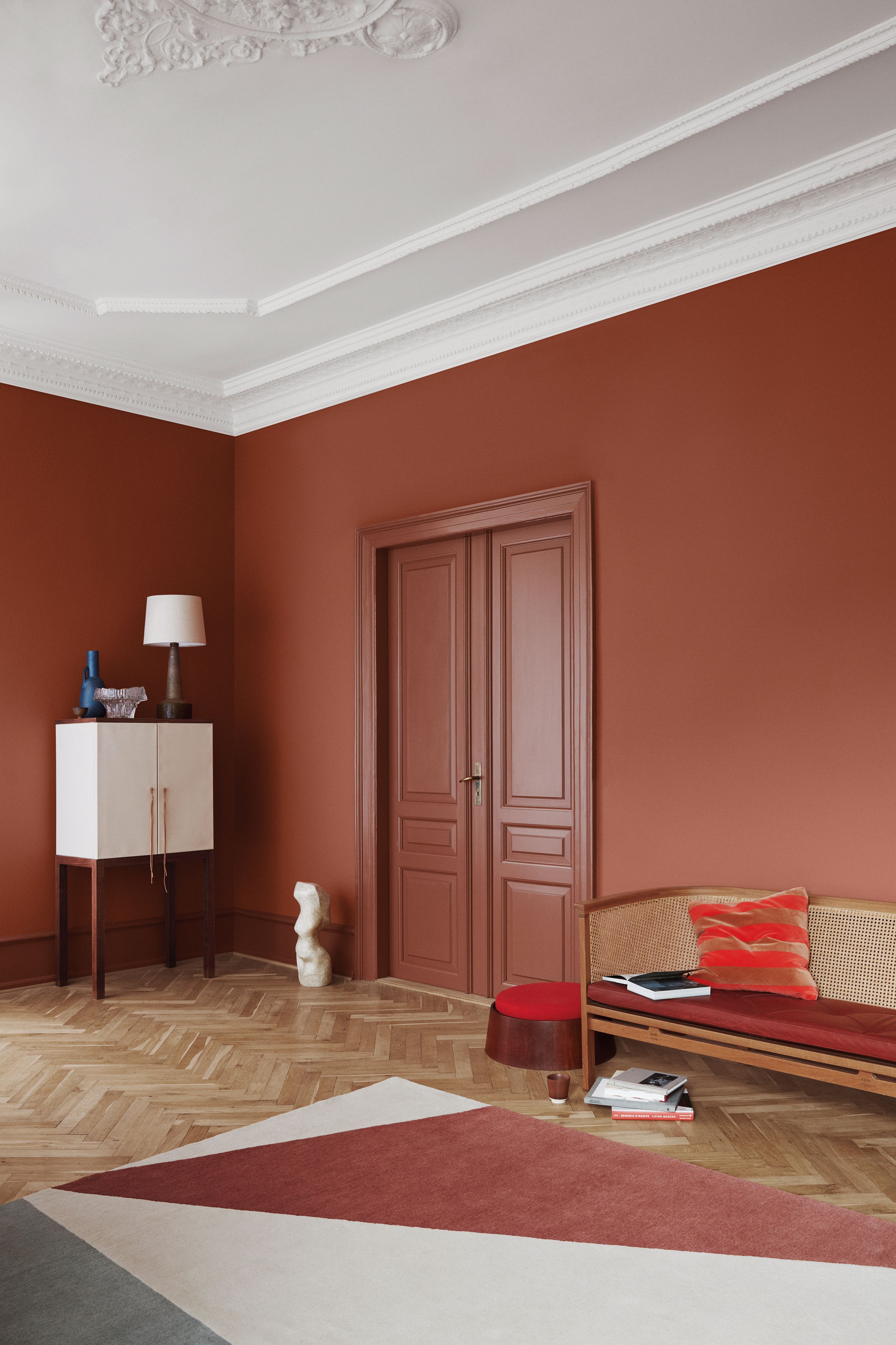A room with terra cotta walls, a white cabinet, a wicker bench, and a geometric rug on a herringbone floor.