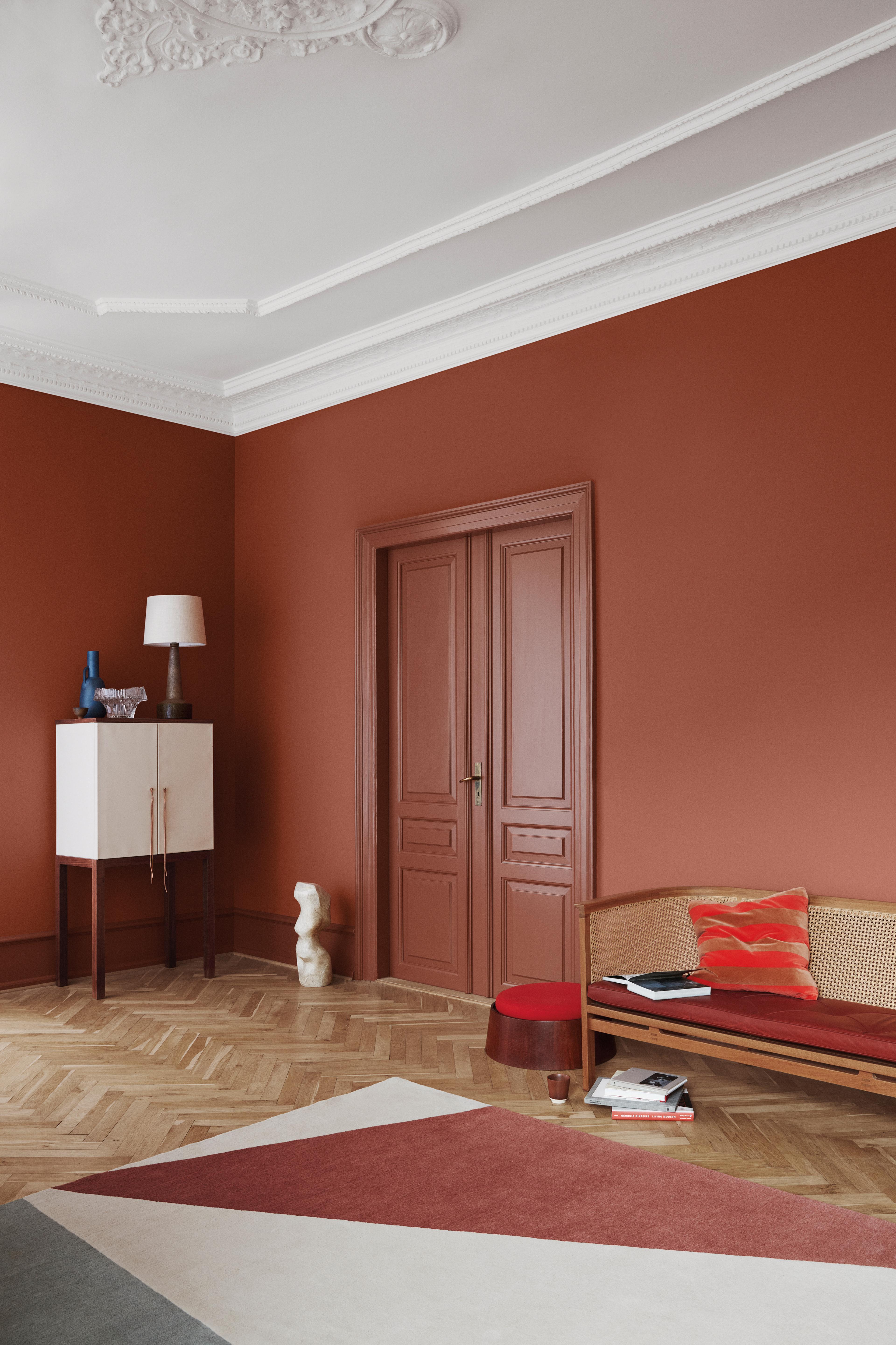 A room with terra cotta walls, a white cabinet, a wicker bench, and a geometric rug on a herringbone floor.