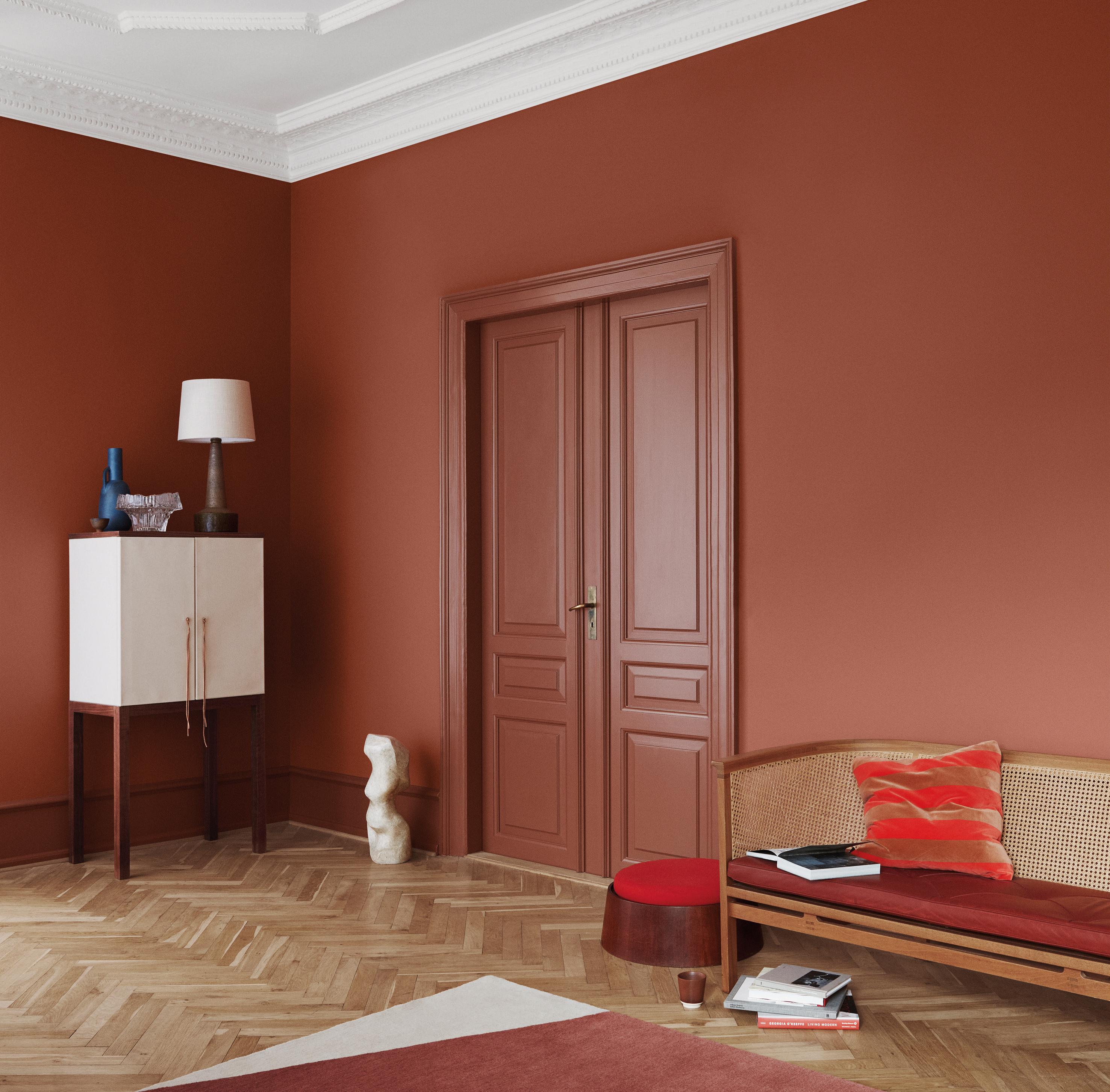 A room with terra cotta walls, a white cabinet, a wicker bench, and a geometric rug on a herringbone floor.