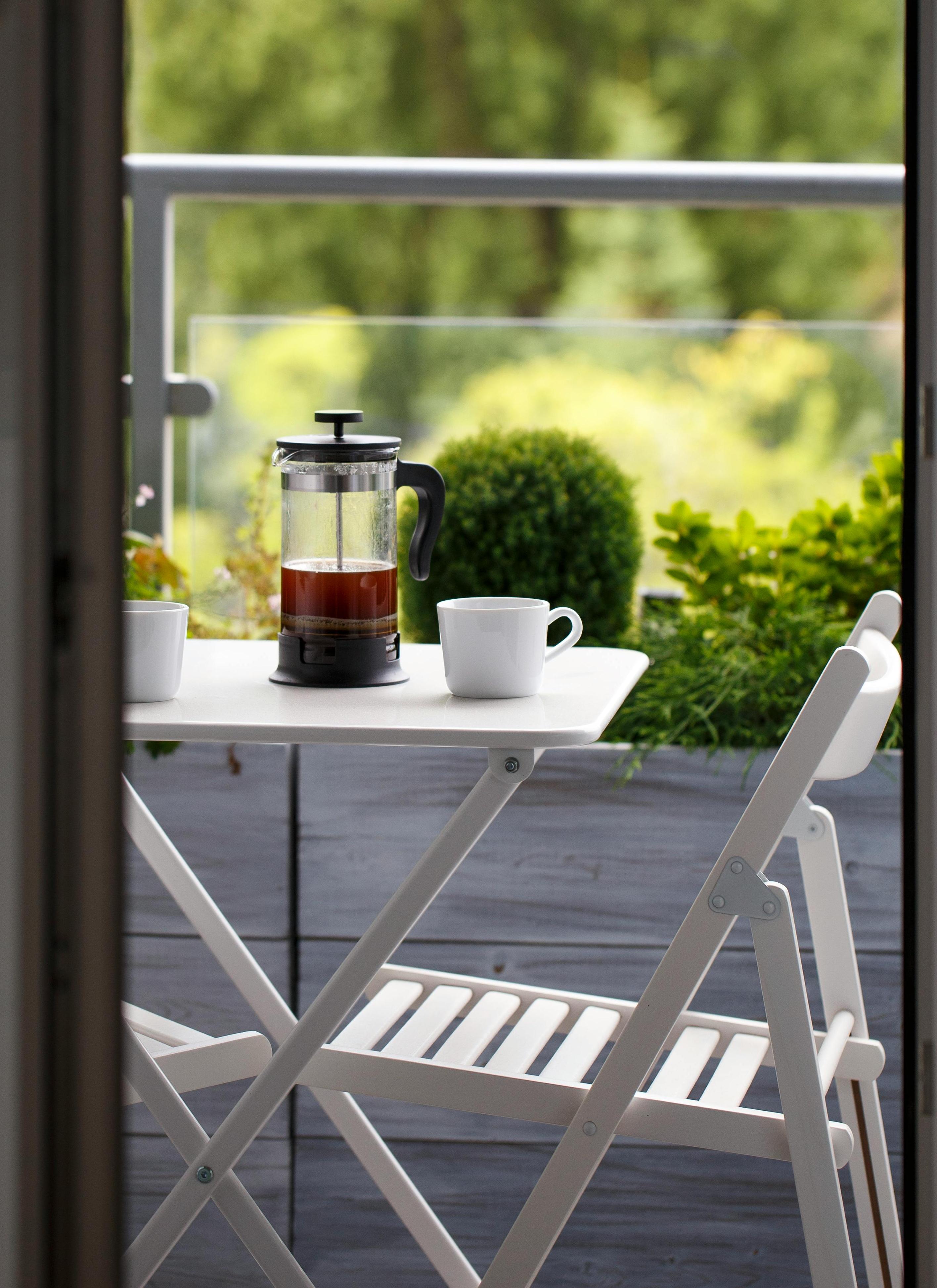 A french press with coffee and two white mugs on a small white balcony table overlooking green foliage.